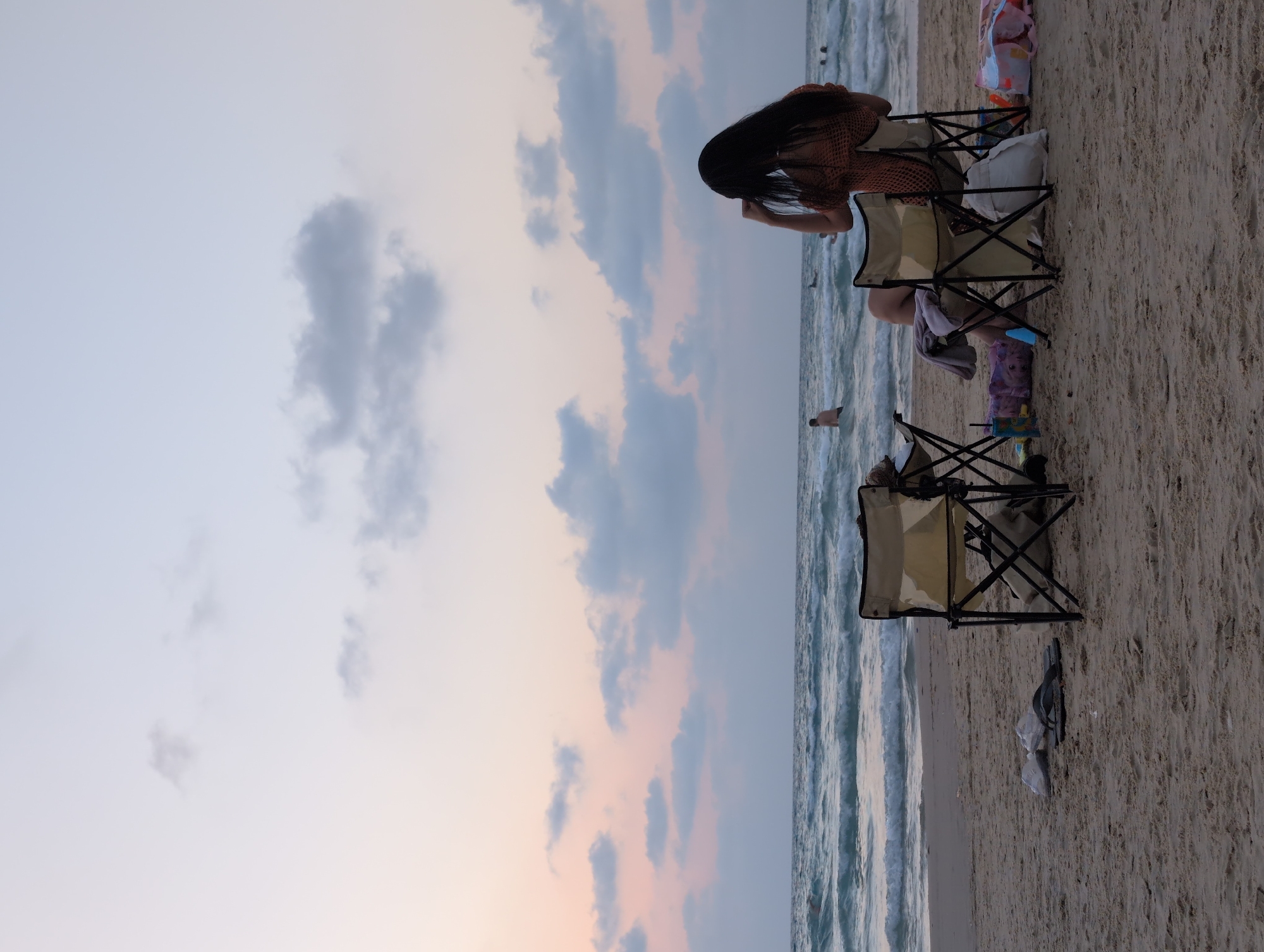 The image depicts a serene evening scene on a beach in Bat Yam, Israel. Two folding beach chairs are set up on the sand, facing the ocean. A woman with long dark hair is seated in the right chair, wearing an orange patterned top and facing away from the camera, appearing to be adjusting her hair. In the left chair, a person is partially visible, with a towel draped over the chair. Scattered on the sand are various beach items including flip-flops, a bag, and a child's toy. The ocean waves gently roll onto the shore under a sky with soft pink and blue hues, indicating either sunrise or sunset. Several people are visible in the water in the distance.