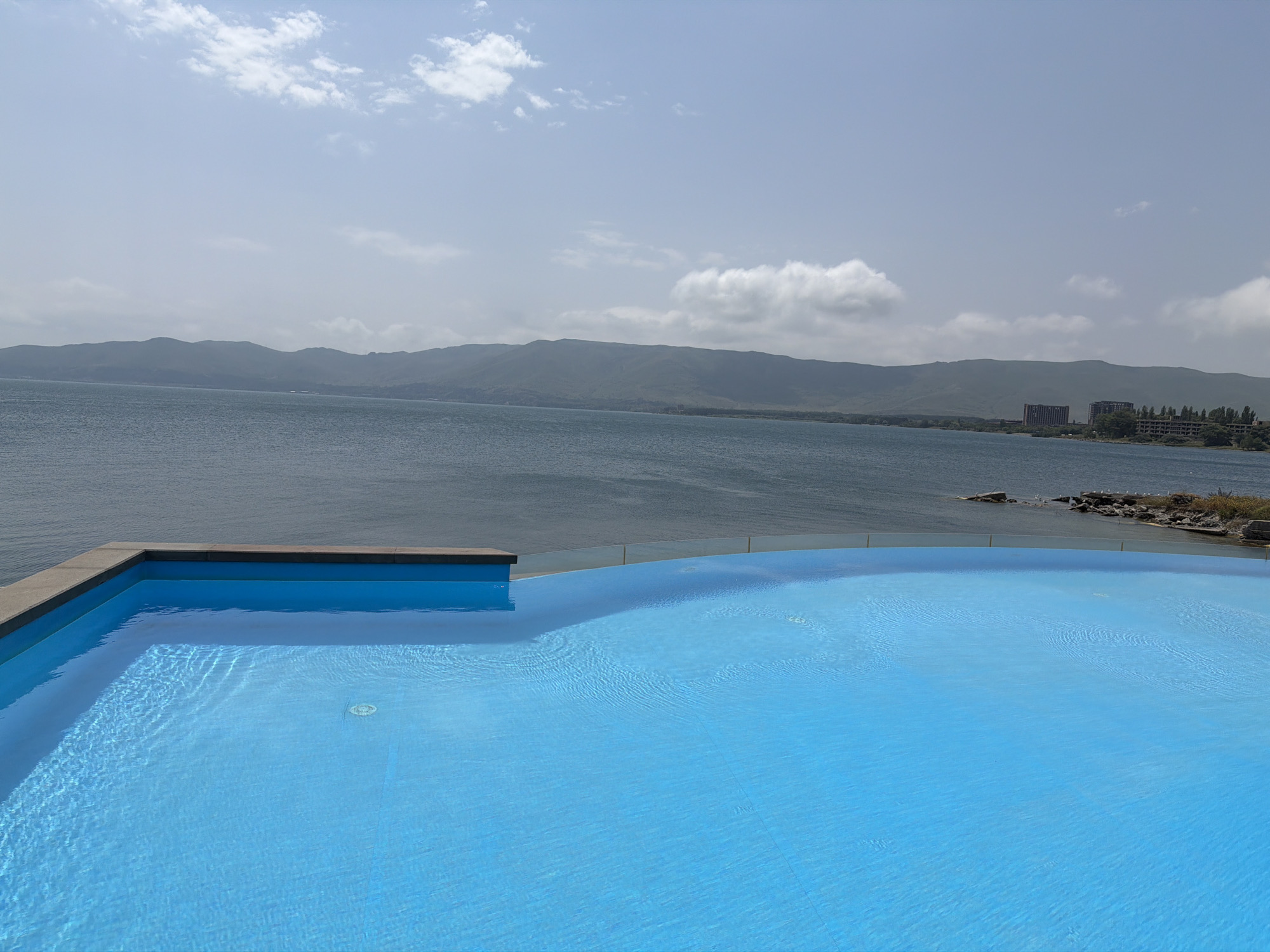 A wide shot captures an outdoor swimming pool with bright blue water, seemingly part of an infinity pool. The pool's edge, made of grey stone, is visible on the left. Beyond the pool, a vast body of water stretches towards a distant shoreline characterized by rolling green hills and mountains. Several buildings, including a tall, dark structure and a longer, low-rise building with trees, are visible along the shore. The sky above is a pale blue with scattered white clouds. The scene appears to be a serene landscape overlooking a large lake or sea, with the pool providing a foreground element.
