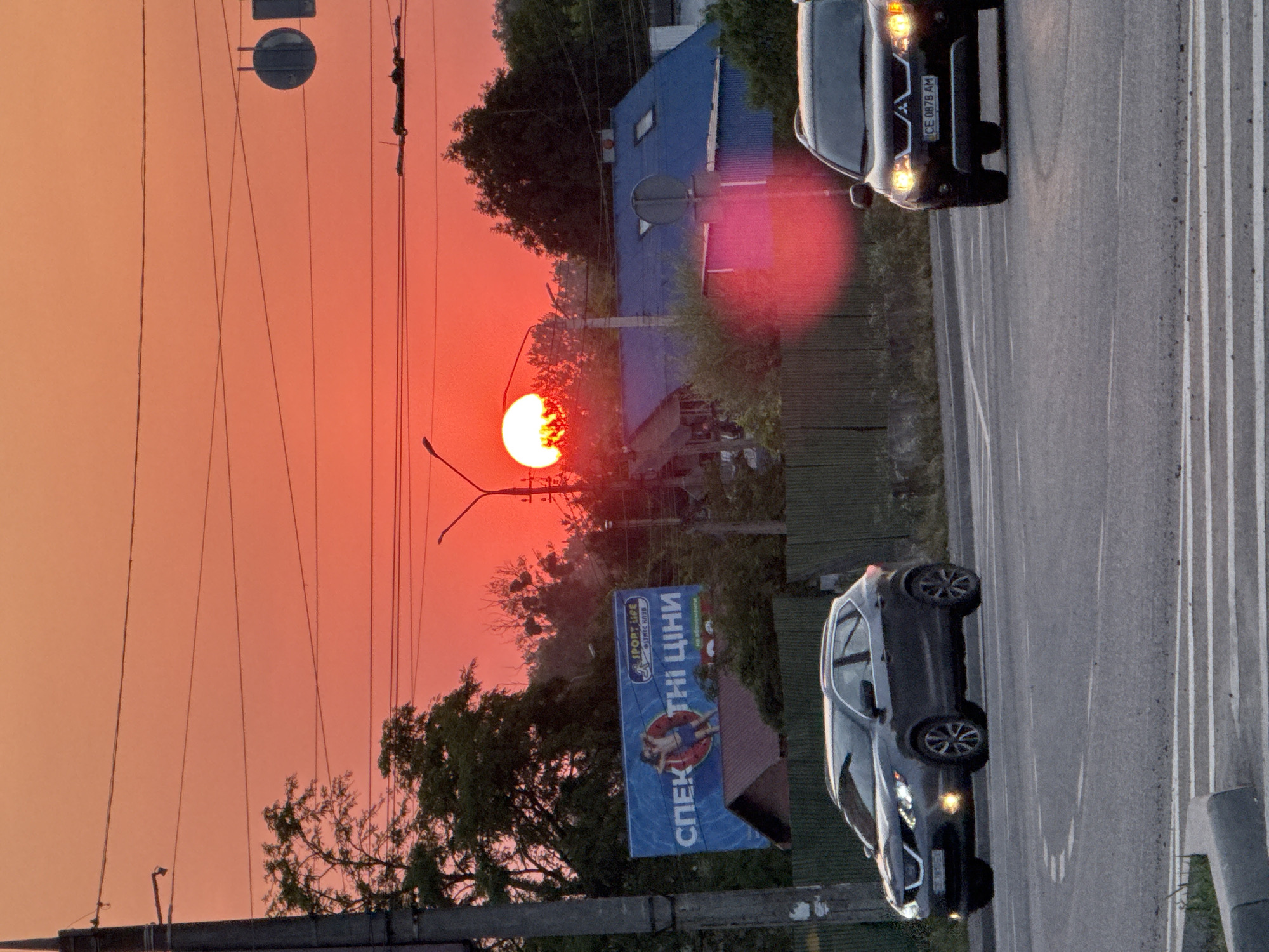 The image captures a vibrant sunset over a street scene in Chernivtsi, Ukraine. The sky is a brilliant orange-red, with the sun partially obscured by trees and a billboard. The billboard features an advertisement for "SPORT LIFE" fitness club with text in Ukrainian. Two cars are visible on the road; a dark SUV on the left and a dark crossover on the right, both appearing to be in motion. A power pole and overhead wires are present on the left side of the frame, and a blue-roofed building with attic windows is situated in the background. The road surface shows lane markings and a pedestrian crossing. The overall impression is an urban setting at dusk, illuminated by the dramatic colors of the setting sun.