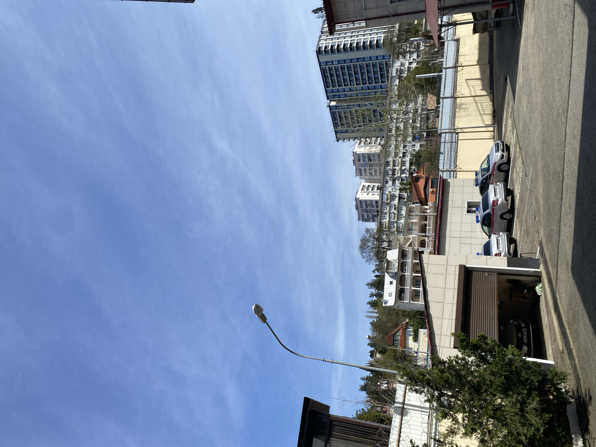 The image shows a street scene in Sochi, Russia. A row of buildings of various heights and architectural styles lines one side of the street. Tall apartment buildings are visible in the background. A parking area is alongside the buildings, with several cars parked. A street lamp stands on the opposite side of the street against a backdrop of a bright blue sky. The image seems to have been taken from a slightly elevated position.