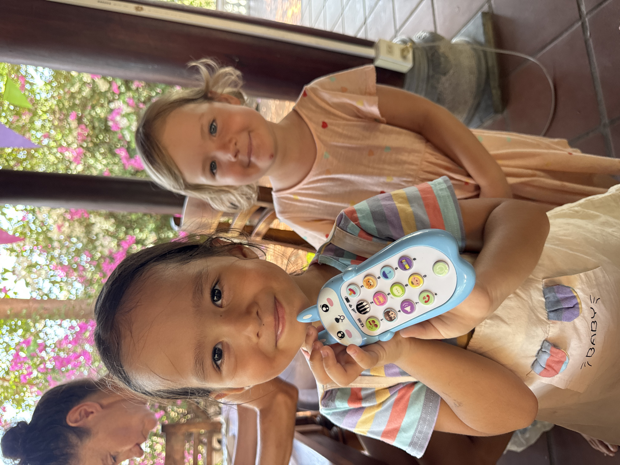 This image, captured in Mũi Né, Vietnam, portrays two young girls in an open-air, tropical setting during the daytime. The bright natural light illuminates the scene, suggesting sunny weather.
In the foreground, a girl with dark hair and dark eyes looks directly at the camera with a gentle smile. She is holding a light blue toy phone shaped like an animal, possibly a bunny or cat. The phone features various colorful circular buttons with icons such as a camera, music notes, an umbrella, an animal face, a phone receiver, a person's profile, a volume symbol, and numbers 1, 2, and 3. The toy phone also displays "12:00". She wears a striped t-shirt in colors including blue, pink, orange, and white, underneath a light-colored apron or dress that has the text "/BABY" written on it.
Slightly behind her and to the right, another young girl with blonde hair styled in pigtails and blue eyes is also smiling at the camera. She is wearing a light pink dress decorated with small heart patterns. Both girls appear happy and engaged. A portion of an adult woman with dark hair tied back is visible in the lower left, looking downwards.
The background showcases lush green foliage punctuated by vibrant pink bougainvillea flowers, typical of a tropical environment. Wooden beams and a tiled floor frame the scene, hinting at a covered patio or open-air dining area. The overall atmosphere is cheerful and relaxed.