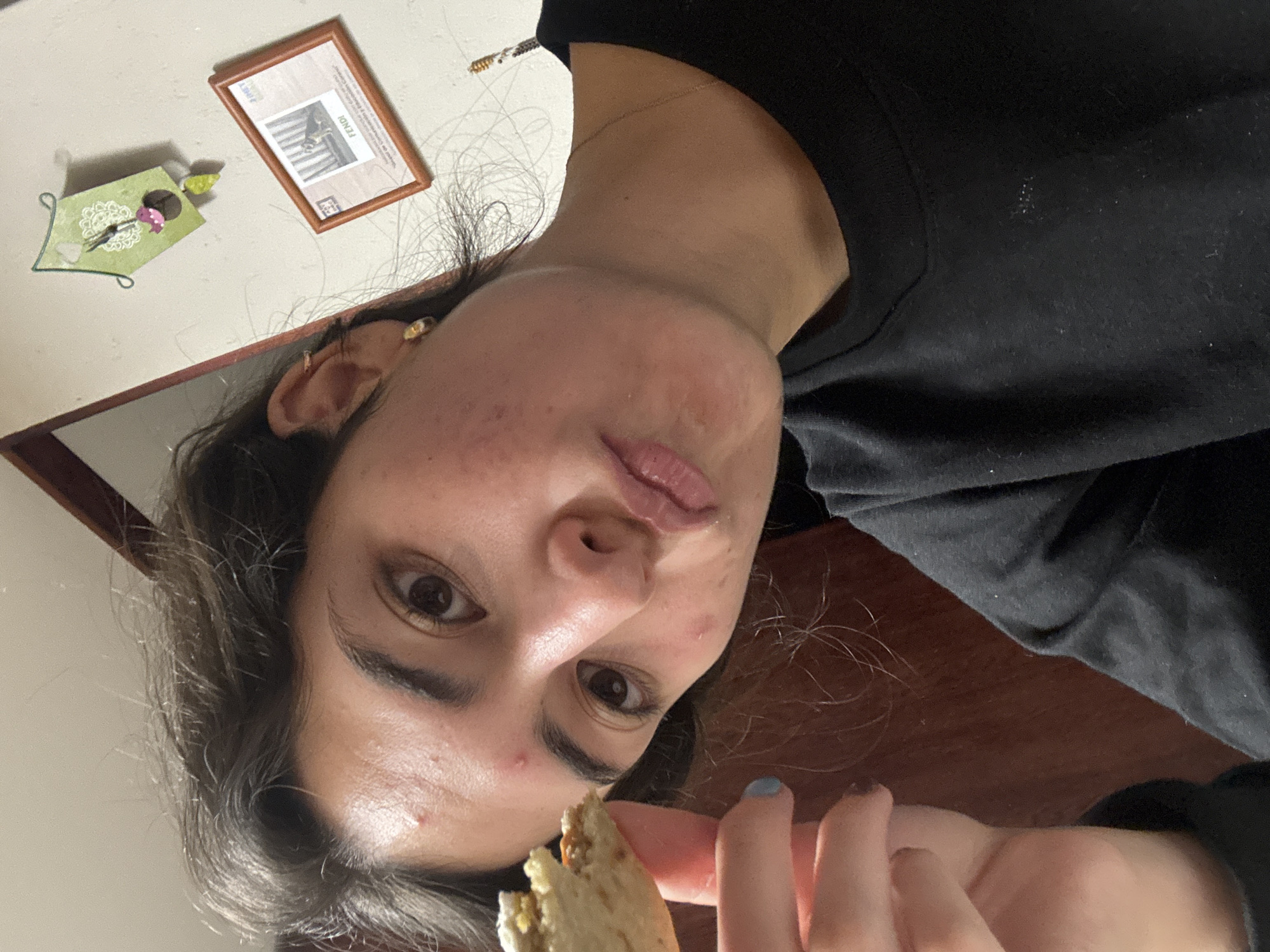 A young woman with dark hair and fair skin is holding a piece of food, possibly bread, near her face. She is wearing a black shirt. Behind her, on a light-colored wall, are a framed picture and a decorative wall hanging. The room has a dark wood surface visible. No specific location in Puebla, Mexico can be determined from this image.