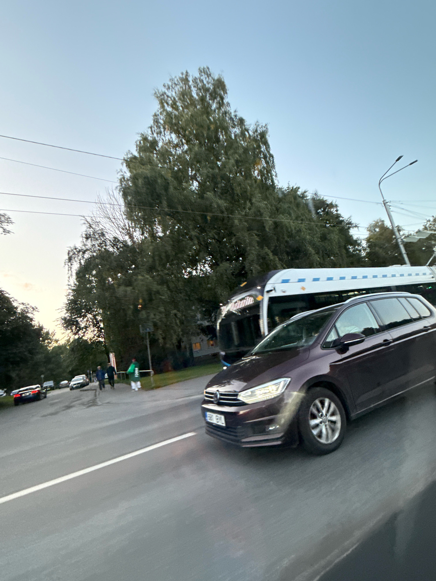 This is a wide-angle, eye-level shot taken from a moving vehicle looking forward and slightly to the right. The scene is a city street in Tallinn, Estonia, likely in the late afternoon or early evening, as indicated by the soft, diffused light and the pale sky.
In the foreground, a dark purple station wagon is the primary focus, captured in motion. Its headlights are on, and the car is moving to the right. The road surface is asphalt with a white lane marking.
Further down the road, a tram is visible. It's white with blue stripes and appears to be stopped or moving slowly. Behind the tram, a dense line of trees lines the street, some with green foliage and others appearing bare or autumnal.
Several pedestrians are walking on the sidewalk to the left of the tram and the trees. One person in the foreground on the sidewalk is wearing a bright green jacket. Other individuals are dressed in darker clothing.
In the background, more trees and some buildings with illuminated windows are visible. A street light with two heads is visible on the right side of the frame, silhouetted against the sky. Several other cars are parked or moving on the street in the distance. Power lines are also visible stretching across the upper part of the frame.