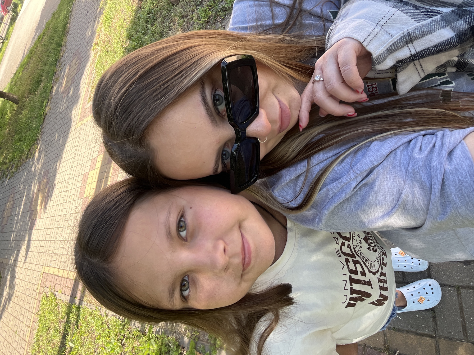 The image captures two individuals, both appearing female, in a selfie-style photograph taken outdoors in the city of Novopokrovskaya, Russia, on a sunny day. The individual on the left is younger, with long brown hair, green eyes, and a small mole on her right cheek, wearing a cream-colored t-shirt with a brown "UNIVERSITY OF WISCONSIN-WHITEWATER" graphic, denim shorts, and white Crocs with orange charms. She looks directly at the camera with a subtle smile. The individual on the right, appearing older, has long brown hair with blonde highlights, wears black rectangular sunglasses, a silver septum piercing, and a silver ring on her left ring finger. Her left hand, with red painted nails, rests near her chin. She is dressed in a grey long-sleeved shirt, partially covered by a grey and white plaid overshirt. She looks over the top of her sunglasses directly at the camera. The background consists of a paved walkway composed of interlocking brown and beige bricks, bordered by green grass and sparse vegetation. In the distant background, a road with a red vehicle is visible.