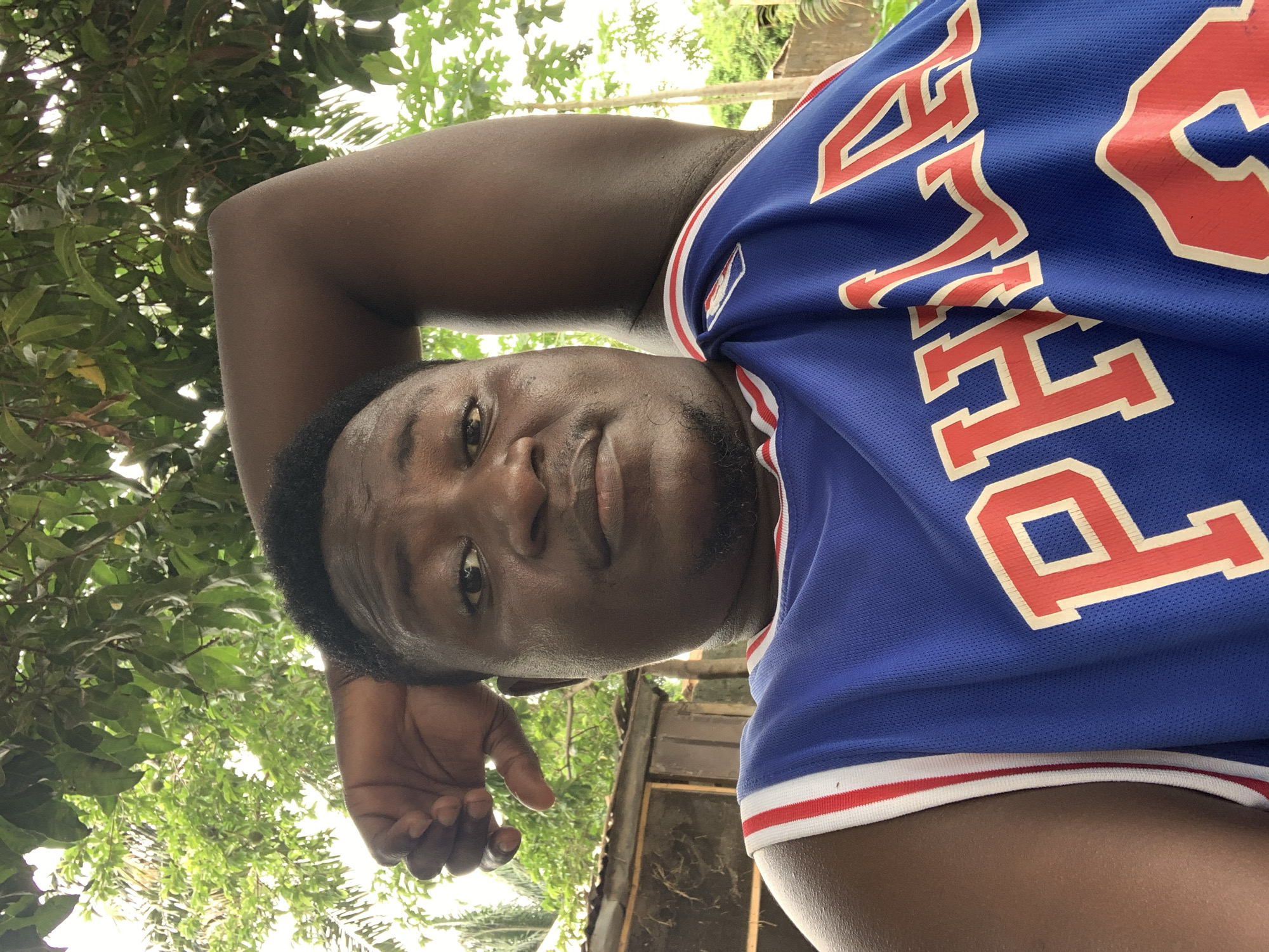The image is a portrait of an adult male, likely a selfie, taken outdoors. The subject, who has dark skin, is positioned slightly off-center and looks directly at the camera with a neutral, calm expression, possibly a faint smile. He has short dark hair and facial hair, appearing as a beard or stubble. His right arm is bent, with his hand resting on or near his head.
He is wearing a blue athletic jersey, characteristic of a basketball uniform, featuring prominent red letters outlined in white. The visible text on the jersey clearly spells "PHILA", indicative of a Philadelphia sports team, most likely the Philadelphia 76ers. The jersey also has a distinct red and white striped trim along the bottom edge and sleeves.
The background is filled with lush green foliage from trees or bushes, suggesting an outdoor, natural environment. Bright, natural light filters through the leaves, indicating it is daytime with clear weather. Some darker, possibly wooden structures are faintly visible behind the man on the right side of the frame, amidst the greenery. The setting is consistent with an outdoor area in Akwanga, Nigeria, with its abundant vegetation.