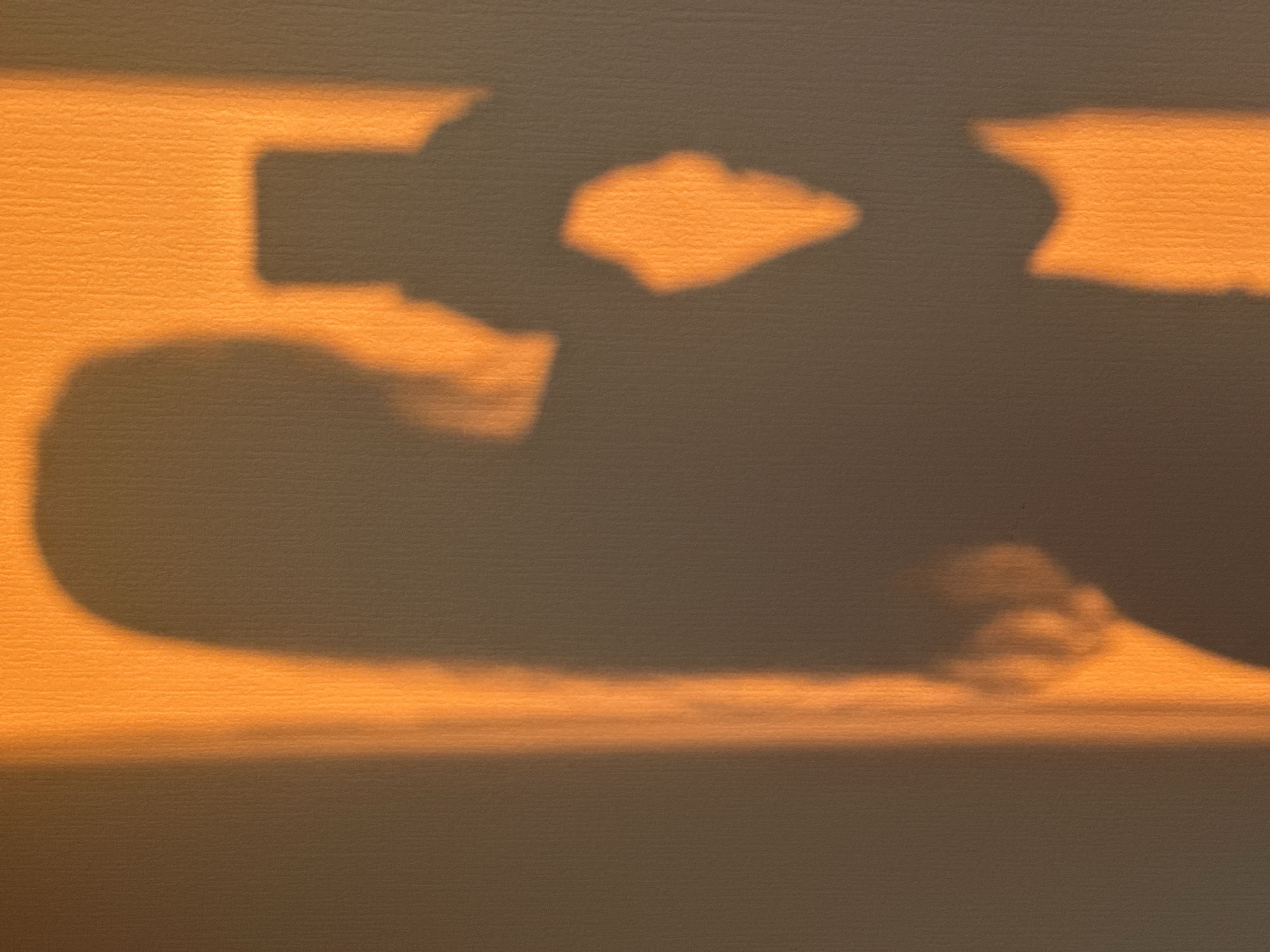 A shadow of a person holding a smartphone is cast on a textured wall under an orange light. The shadow depicts the person from the chest up, with their hair visible. The smartphone is held in front of the person, suggesting they are taking a selfie or recording a video. The light source, likely from a window or lamp, creates a warm, golden hue across the wall and the shadow. The wall's texture is apparent, with subtle vertical ridges. The environment appears to be indoors.
