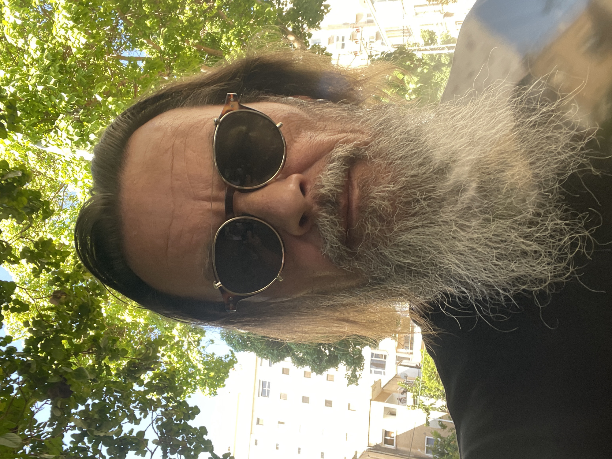 The image is a selfie taken from a low angle, showing a man with long, light brown hair and a full, greyish-white beard. He is wearing round, tortoiseshell-rimmed sunglasses. The background features lush green trees with sunlight filtering through the leaves, and in the distance, several multi-story buildings are visible. One building is a light beige color with a grid of windows, and another to its right appears to be white. The overall setting suggests an outdoor urban environment, likely in Netanya, Israel, given the architectural style of the buildings and the Mediterranean foliage. The man's expression is neutral, and he appears to be looking directly at the camera. The lighting is bright, indicating a sunny day.