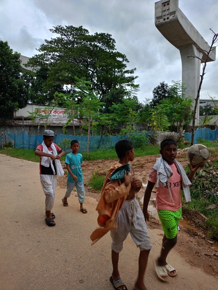 The image depicts four male juveniles walking on an unpaved dirt road in Sujabad, Bangladesh, under an overcast sky. The road surface is brown earth with some wet patches on the right.
From left to right:
1. A juvenile wearing a white skullcap, a red short-sleeved shirt, and white loose-fitting trousers. He is positioned in the foreground, adjusting a white cloth draped over his shoulders. His footwear consists of dark slip-on sandals.
2. Behind the first juvenile, a second juvenile wears a light blue short-sleeved t-shirt with a "Spotify" logo and light blue trousers. He walks in dark slip-on sandals.
3. A third juvenile, partially obscured, wears a tan long-sleeved shirt draped over his left shoulder and light grey loose shorts or trousers. An orange object, possibly a fruit, is partially visible in his left hand. He wears striped slip-on sandals.
4. On the far right, a fourth juvenile wears a pink short-sleeved t-shirt with "XBT" text, bright green patterned shorts, and light-colored slip-on sandals. He holds a deflated, worn, patched soccer ball in his left hand, and a white cloth is draped over his right shoulder.
In the mid-ground, a blue mesh fence separates the road from an area of dense green vegetation. Behind this vegetation, a white building with a sign featuring Bengali script, potentially reading "Kalyanpur College," is visible. To the far right, a tall, grey concrete pillar, indicative of ongoing bridge or elevated road construction, extends vertically with exposed rebar points at its top. A large, mature tree with dense green foliage dominates the upper central background. The sky is uniformly cloudy.