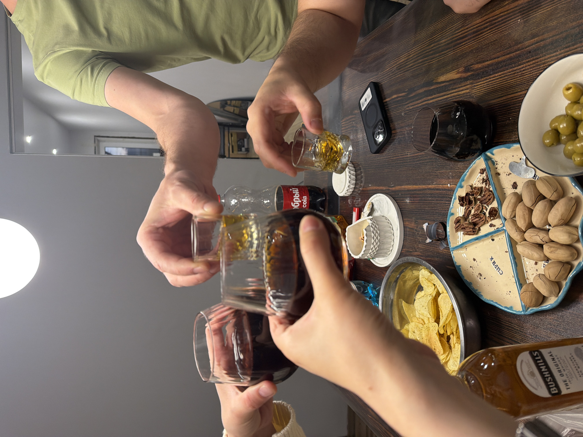 A high-angle shot shows a group of people clinking glasses at a table indoors. The table is made of dark-stained wood and is laden with various items. Hands hold three glasses, one containing a clear, amber liquid, one with a darker amber liquid, and the third with a dark red liquid. Adjacent to the glasses, there is a small shot glass with a clear, amber liquid. A bottle of Coca-Cola is present, along with an empty clear plastic bottle. A black smartphone sits on the table, next to a small white candle in a decorative holder.
The table also features a bowl of yellow potato chips, a divided dish containing nuts and other snacks, and a small plate of green olives. A bottle of Bushmills whiskey is visible along the right edge of the frame.
The people involved are wearing casual clothing, including a light green t-shirt and a cream-colored sweater. The background shows a wall and a diffused light source. The location includes the word "ЧАИК", which, according to the dish, suggests that the place is located in the city of Chaikovsky, Russia.