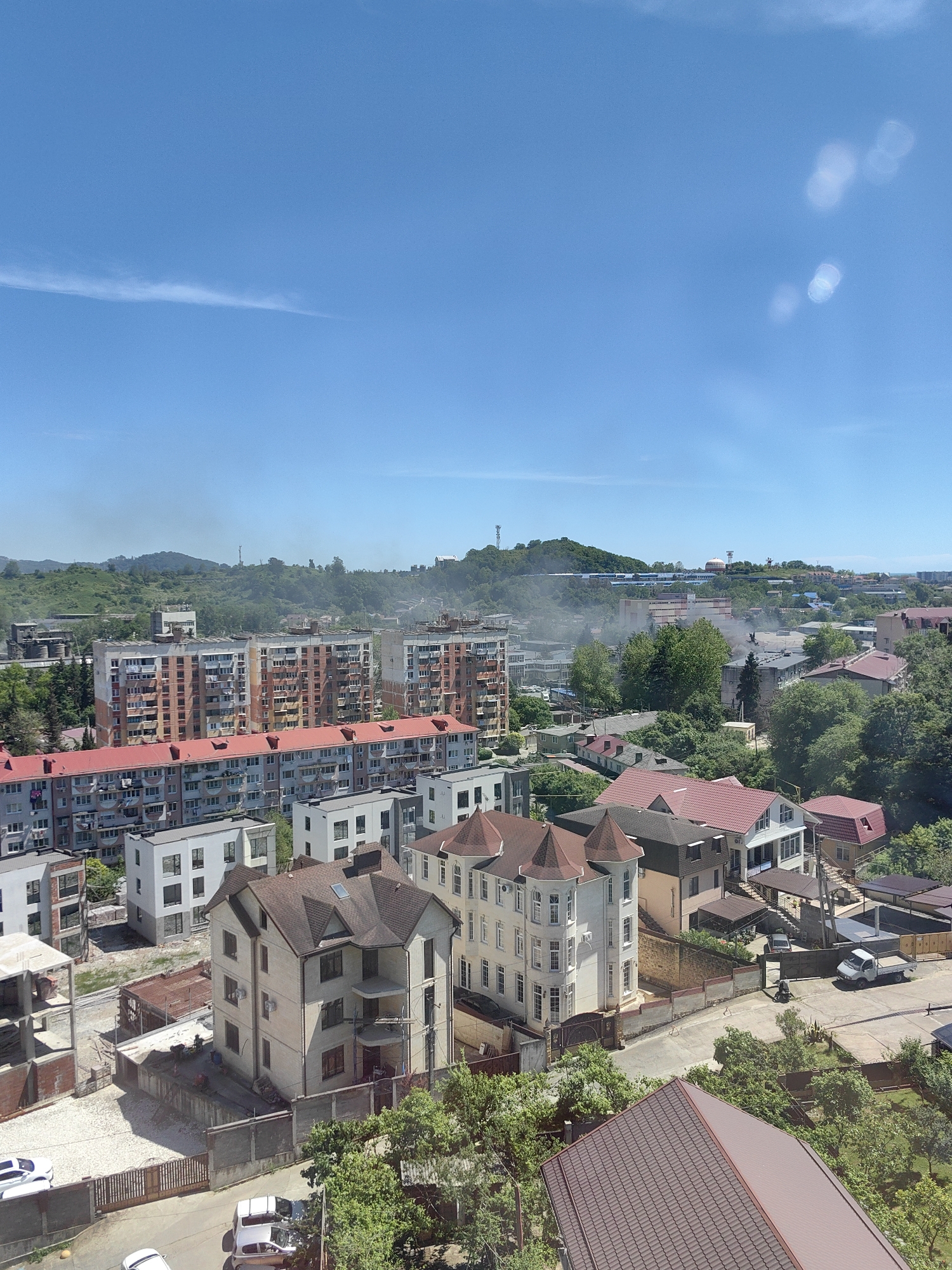 An elevated view captures an urban landscape in Sochi, Russia, under a clear, bright blue sky. The scene is dominated by a mix of residential buildings. In the mid-ground, several large, multi-story apartment blocks, indicative of Soviet-era architecture, feature numerous windows and balconies, some with visible laundry. Surrounding these and in the foreground are numerous detached houses, varying in style from modern designs with clean lines to more ornate structures, often topped with sloped, reddish-brown tiled roofs. Some buildings in the foreground appear to be under construction or renovation.
Hills covered in lush green vegetation form the distant background, with a communication tower visible on one peak. Several plumes of grey-white smoke or haze rise from different points in the mid-ground, suggesting industrial activity or local burning. Cars are parked on visible roads and in driveways. The bright sunlight creates distinct shadows and causes several circular lens flares to appear in the upper right portion of the sky. The time of day is clearly daytime, with sunny weather. No discernible text or individuals are clearly visible.