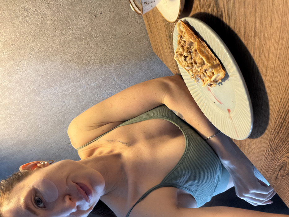 The image displays an indoor scene featuring a woman and a meal on a wooden table, against a textured wall. The woman is positioned on the left side of the frame, looking towards the camera. She has light-colored eyes and full lips, and is wearing light-colored under-eye patches on her cheeks. Her hair is not clearly visible, but a small hoop earring is seen on her left ear. She is wearing a light green, ribbed tank top with thin straps. A faint tattoo of text is visible on her upper chest/collarbone area. On her left wrist, she wears a thin, silver-colored chain bracelet.
To the right of the woman, on a warm-toned wooden surface (likely a table), rests an oval, light-colored plate with a ribbed or fluted edge. On the plate is a piece of food that appears to be a waffle, pancake, or similar pastry, generously topped with a light brown, chunky substance, possibly crushed nuts, a streusel, or a savory filling. A reddish streak or design is also visible on the plate near the food. Further back on the table, to the left of the plate, is a light-colored cup or mug with a handle, sitting on a matching saucer. The cup has small, dark speckles or patterns on its surface.
The background to the upper left of the woman is a textured wall, light grey or beige in color, possibly concrete or a similar material. The lighting in the scene appears to be warm, casting shadows from the objects onto the wooden table. The composition suggests a self-portrait or selfie, taken from a slightly low angle.