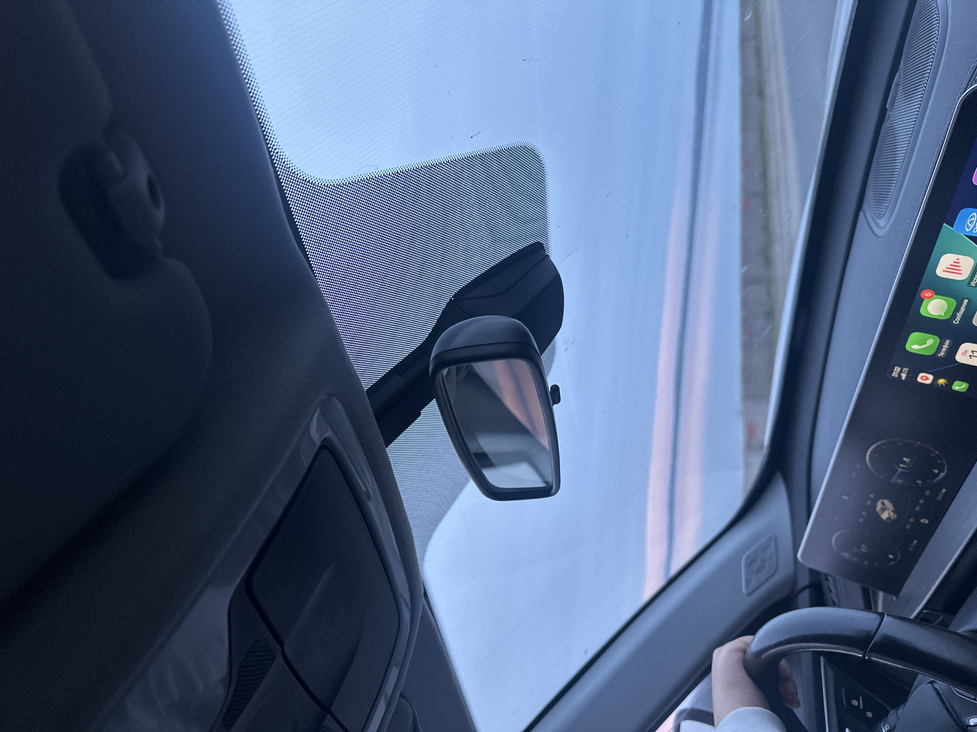 The image shows the interior of a vehicle. A rearview mirror is visible, as well as a section of the dashboard and steering wheel. A hand is gripping the steering wheel. The infotainment screen displays applications. The background outside the windshield shows a blurry street. No specific location in Chaikovsky, Russia, can be determined from the information provided.