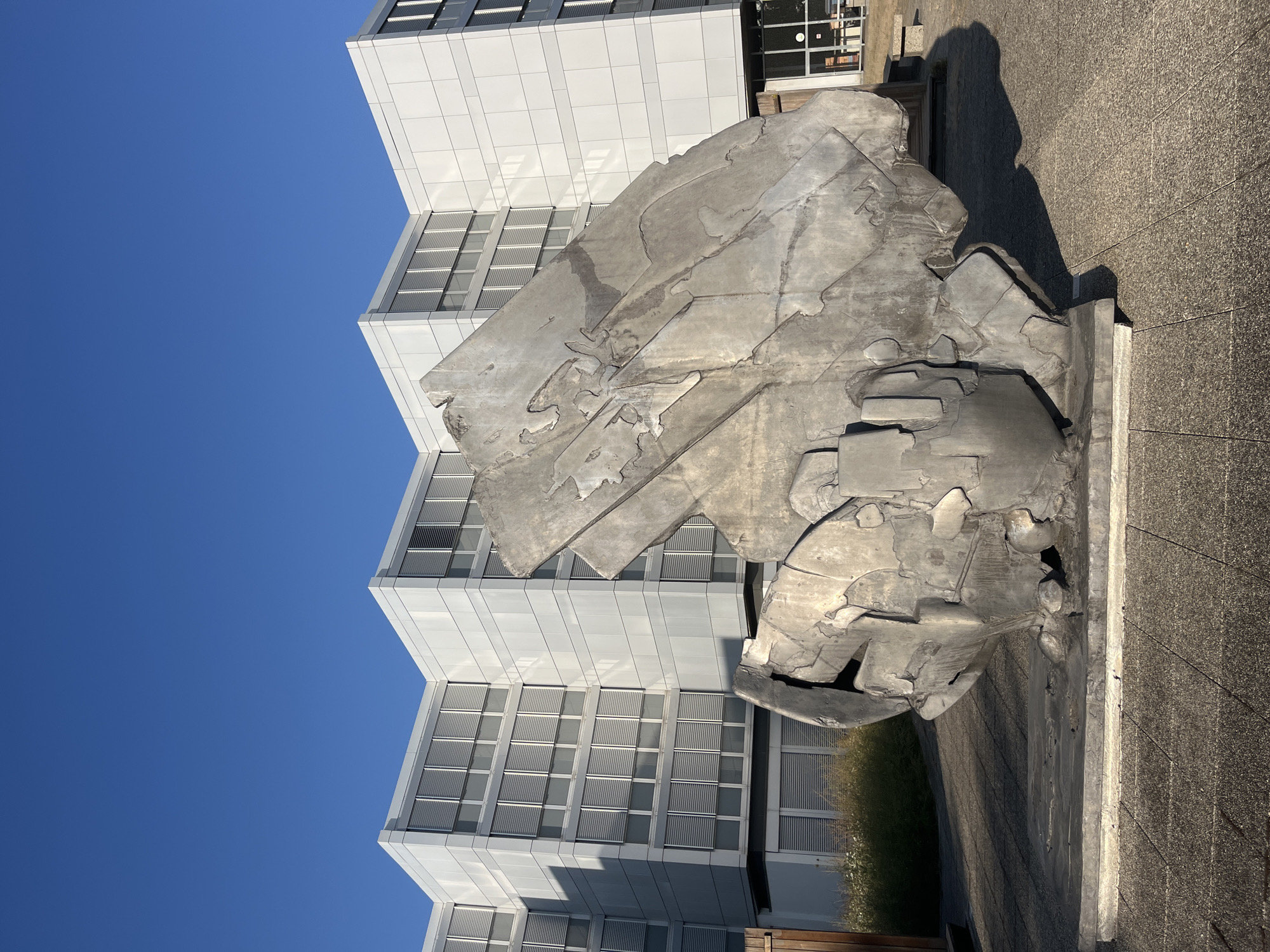 A large, abstract metal sculpture stands in front of a modern, multi-story building under a clear blue sky. The sculpture is composed of irregularly shaped, jagged pieces of metal welded together, giving it a rough, organic appearance. It is situated on a concrete base. The building behind it is clad in light-colored panels and features numerous windows with horizontal blinds. The surrounding area appears to be an outdoor public space with a paved walkway and some low-lying vegetation to the left of the sculpture. The lighting suggests it is daytime, with the sun casting shadows. This scene is located at the CERN Meyrin site, specifically in front of the Main Auditorium building. There are no people visible in the image.