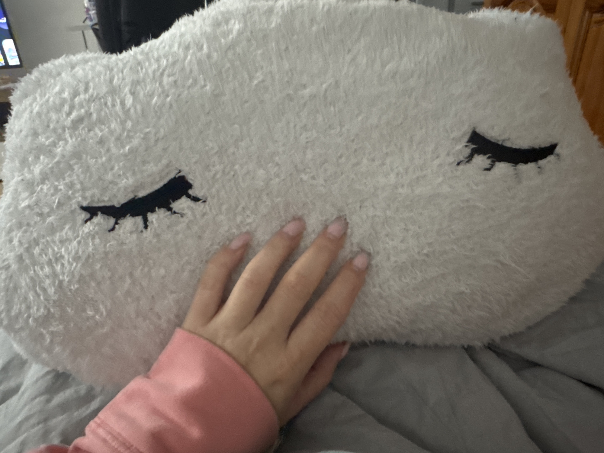 A hand rests on a white, cloud-shaped pillow with stylized closed eyes embroidered in black. The hand is in a pink long-sleeved garment. The pillow is on a gray fabric surface. A partially visible cabinet or wooden furniture is behind the pillow. The image was not taken in Sofia, Bulgaria.