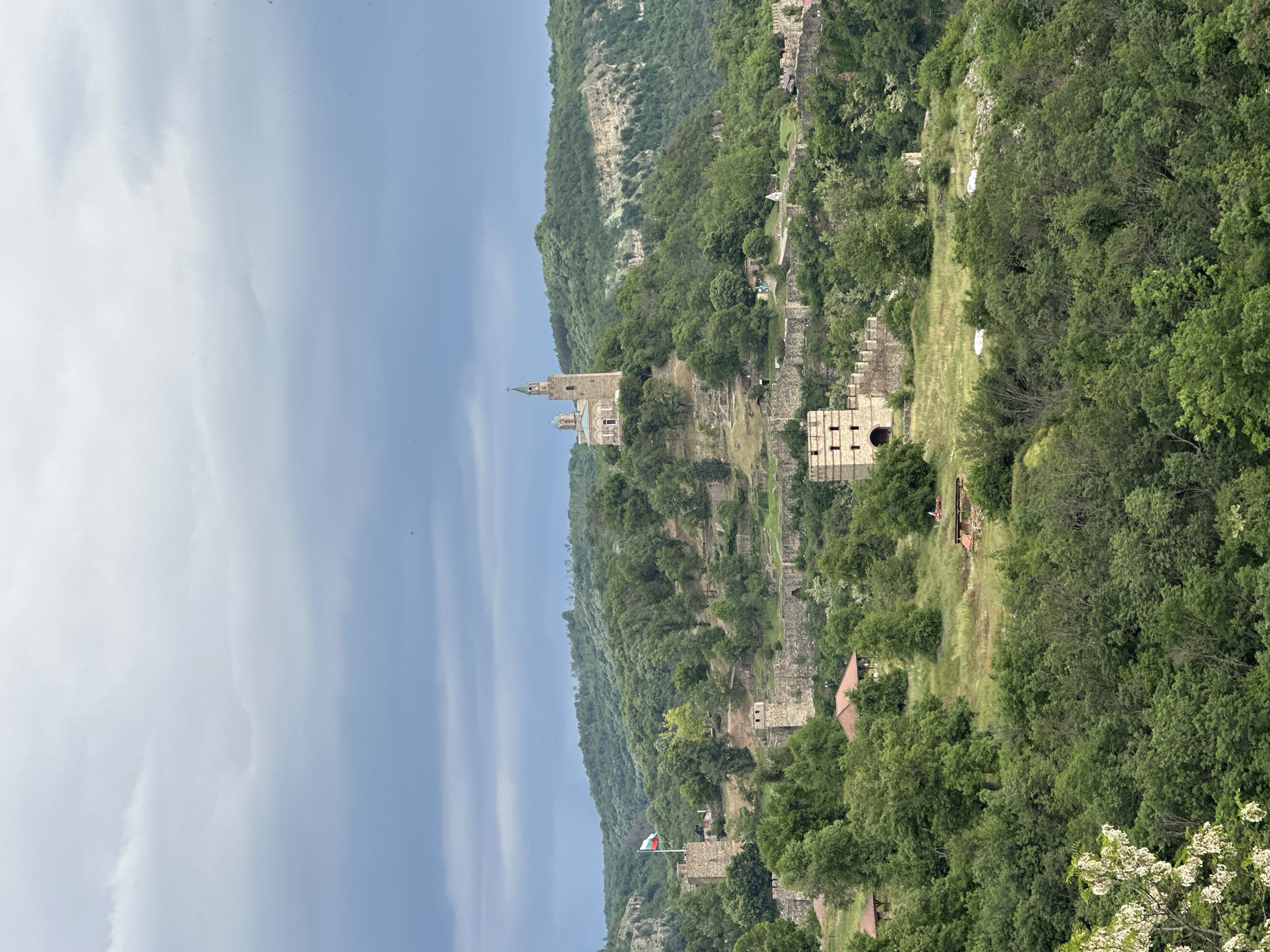 This image shows the Tsarevets Fortress in Veliko Tarnovo, Bulgaria. The fortress is situated on a hill, surrounded by a dense forest. A tall church with a spire is prominently visible at the highest point of the fortress. Parts of the stone walls and ruins of the fortress are visible on the hillside, with the Bulgarian flag flying. The sky is overcast.