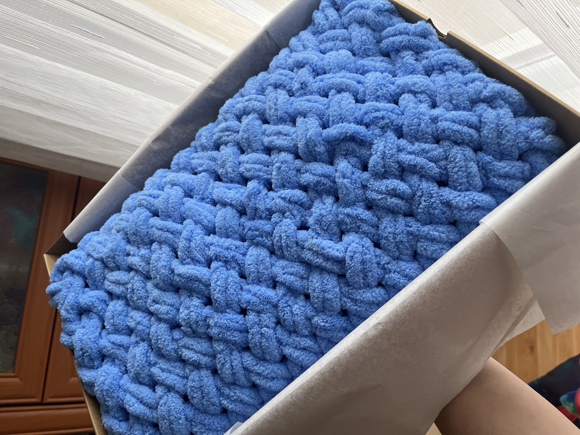 A close-up shot shows a blue, hand-knitted blanket nestled inside a cardboard box lined with white tissue paper. The thick yarn is woven in a repeating pattern. The box is being held by a person whose arm is visible on the right. In the background, part of a brown wooden cabinet is visible, as well as a set of white window blinds. The location is unknown.