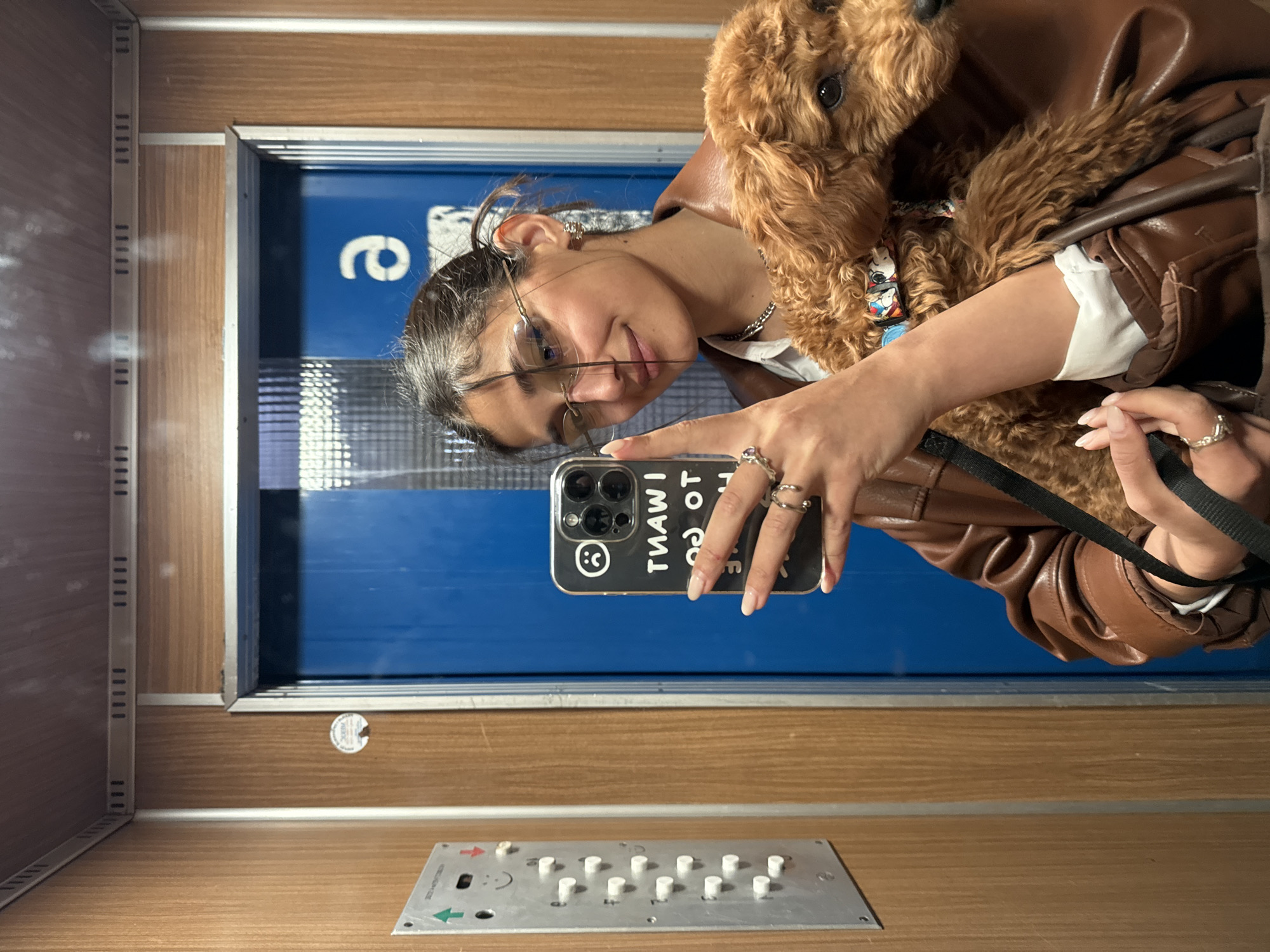 A woman with brown hair and glasses is taking a selfie with her brown poodle in an elevator. She is wearing a brown leather jacket with a furry collar. The elevator has a blue door and wooden paneling. There is a control panel with buttons visible on the floor. The location is the city of Bar, Montenegro.