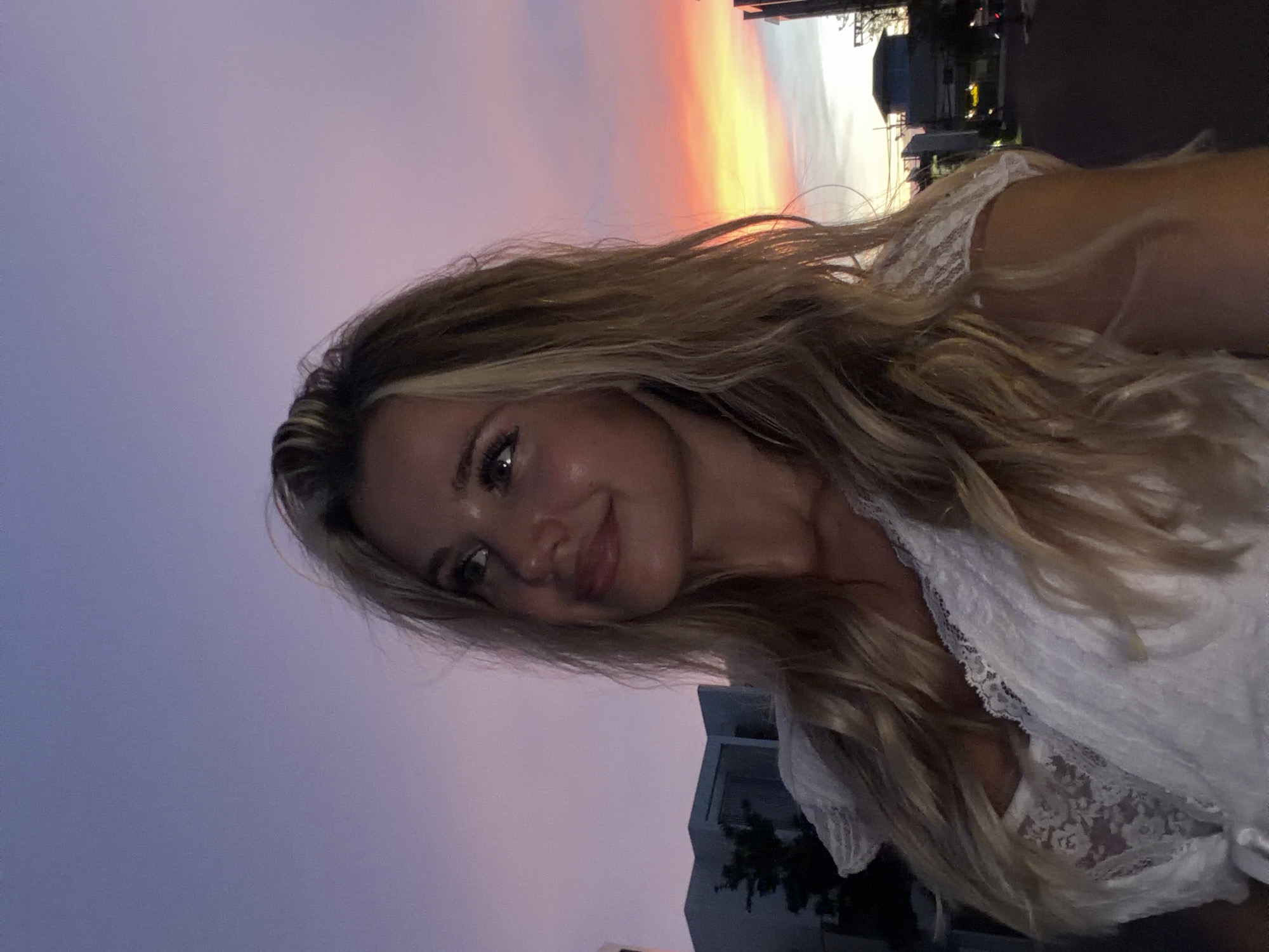 A young Caucasian woman with blonde hair and fair skin is captured in a selfie at dusk. She is wearing a white lace top. Her head is tilted upwards, looking to her left with a slight smile. The background shows a colorful sky with hues of purple, pink, and orange, typical of a sunset. Silhouettes of buildings and trees are visible in the distance, suggesting an urban environment. The specific location of Thep Krasatti, Thailand, is not evident from the visual information in the image.