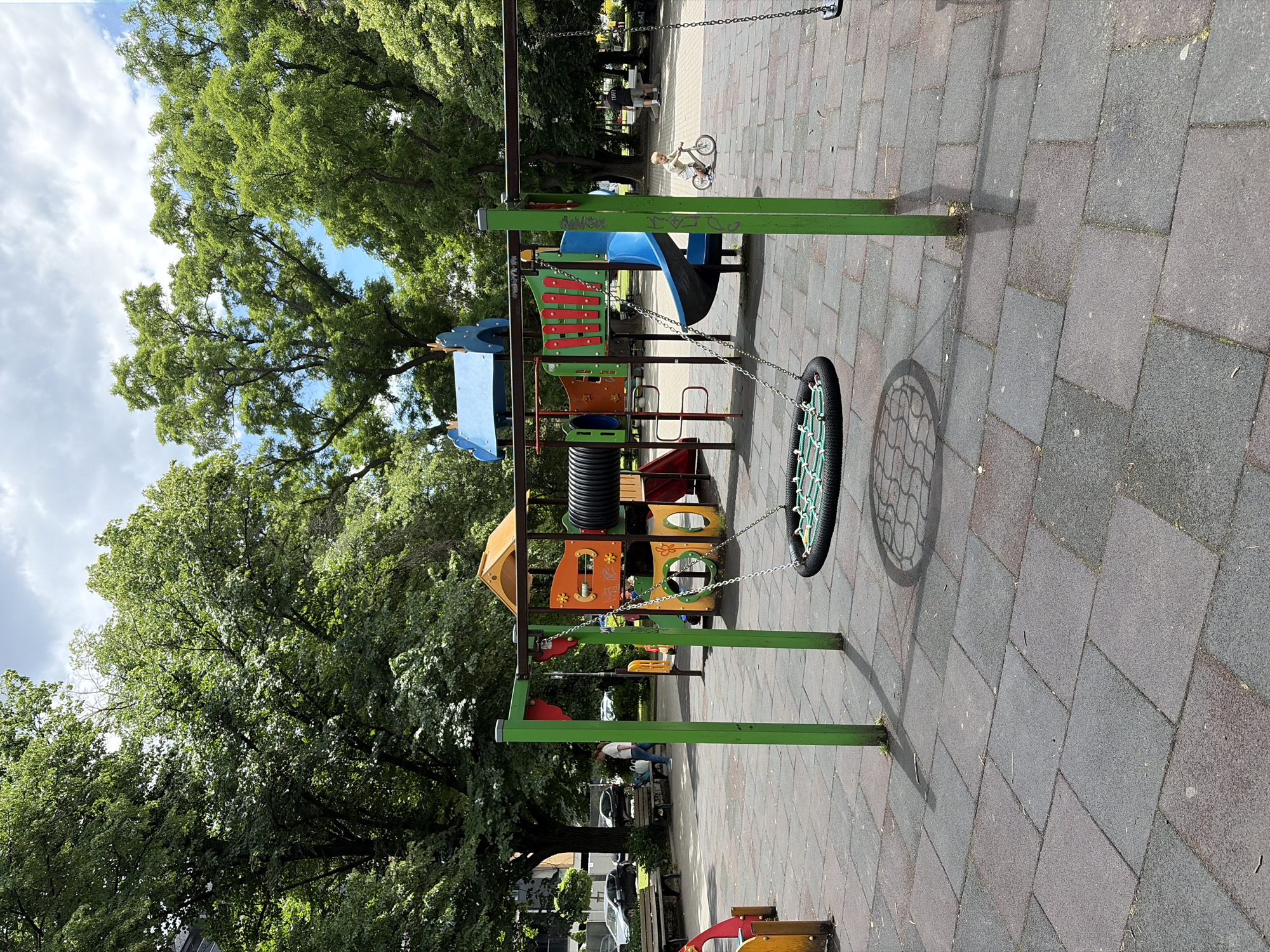 This image depicts an outdoor playground scene in Sofia, Bulgaria, under a partly cloudy sky. In the foreground, a paved area consists of grey and dark grey interlocking rubber tiles. A metal swing set with green posts is prominent, featuring a circular net swing suspended by chains. To the right, a young child on a small bicycle rides across the playground.
The playground structure itself is colorful, with elements of blue, orange, yellow, and red. It includes a slide, tunnels, and climbing features. Behind the playground, mature trees with lush green foliage line the perimeter. In the middle ground, several benches are visible, and a woman in a white shirt and blue pants walks past them. A few cars are parked in the background on a street adjacent to the playground, and a building is partially visible behind the trees on the left. The overall environment suggests a public park or recreational area within the city.