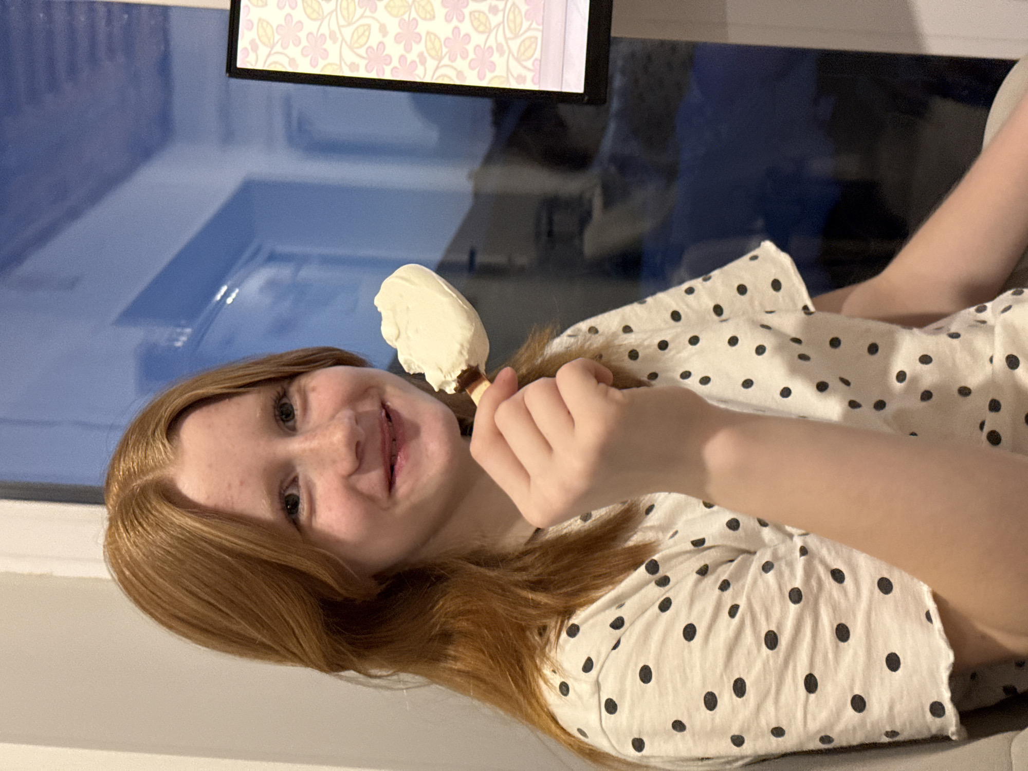 A young, fair-skinned person with red hair is holding a white ice cream bar in their right hand. They are wearing a white shirt with black polka dots. The background includes a window reflecting an interior space with a framed picture or monitor above. The location is unspecified and cannot be determined to be Chaikovsky, Russia from this image.