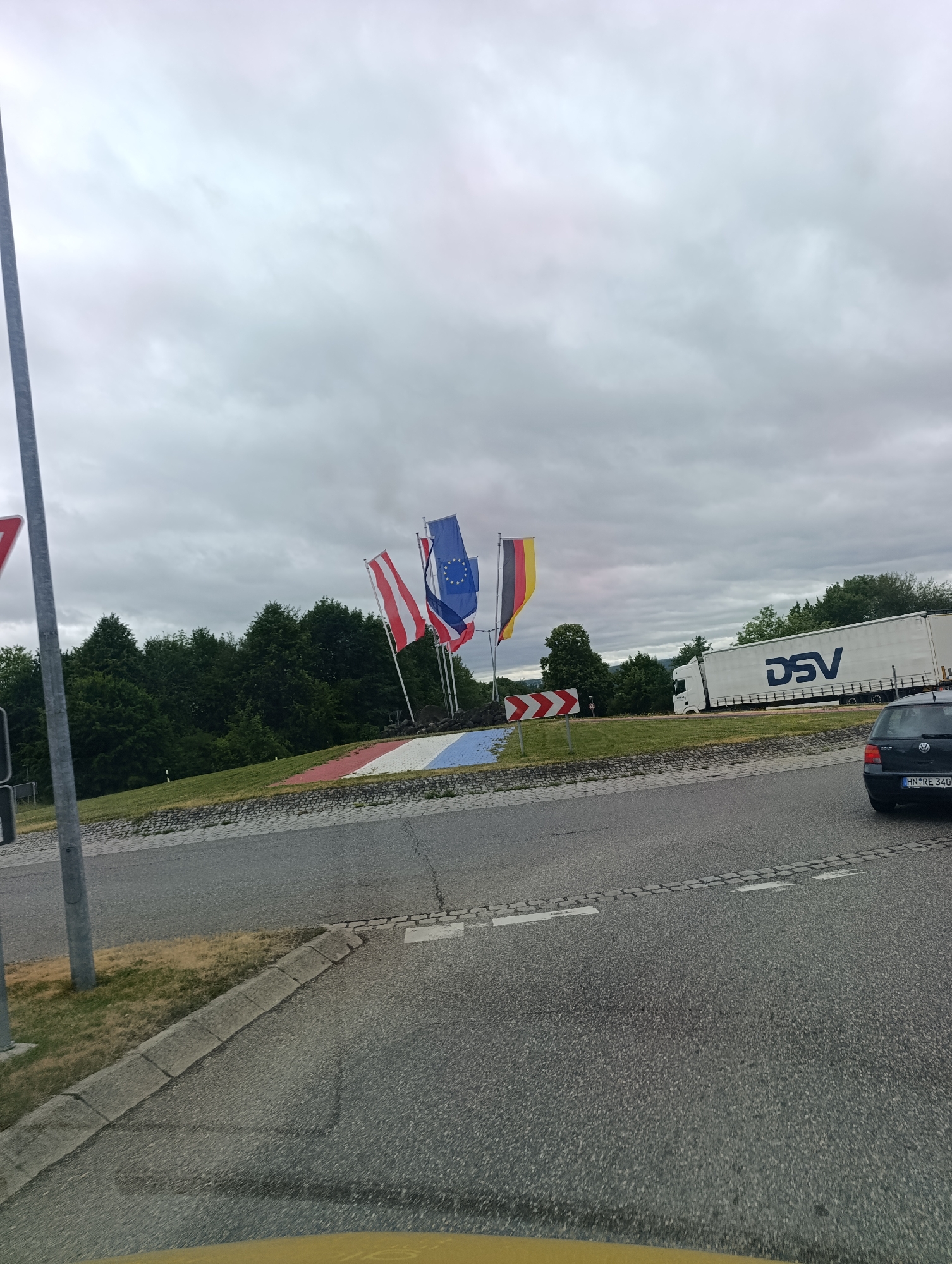 This is an eye-level, medium shot of a roundabout in Sigmaringen, Germany, on a cloudy day. In the center of the roundabout, there are several flags displayed on poles: the Austrian flag, the European Union flag, the French flag, and the German flag. The roundabout itself is decorated with red, white, and blue stripes on a circular grass area. Adjacent to the roundabout, a large white DSV truck is visible driving on the road. A black Volkswagen Golf is parked to the right, facing away from the camera, and its license plate reads "HN*RE 340". The road surface is asphalt with some visible cracks and paving stones along the edge of the roundabout. Trees and vegetation form a backdrop behind the roundabout, and a grey pole with a red and white triangular sign is positioned on the left side of the frame.
