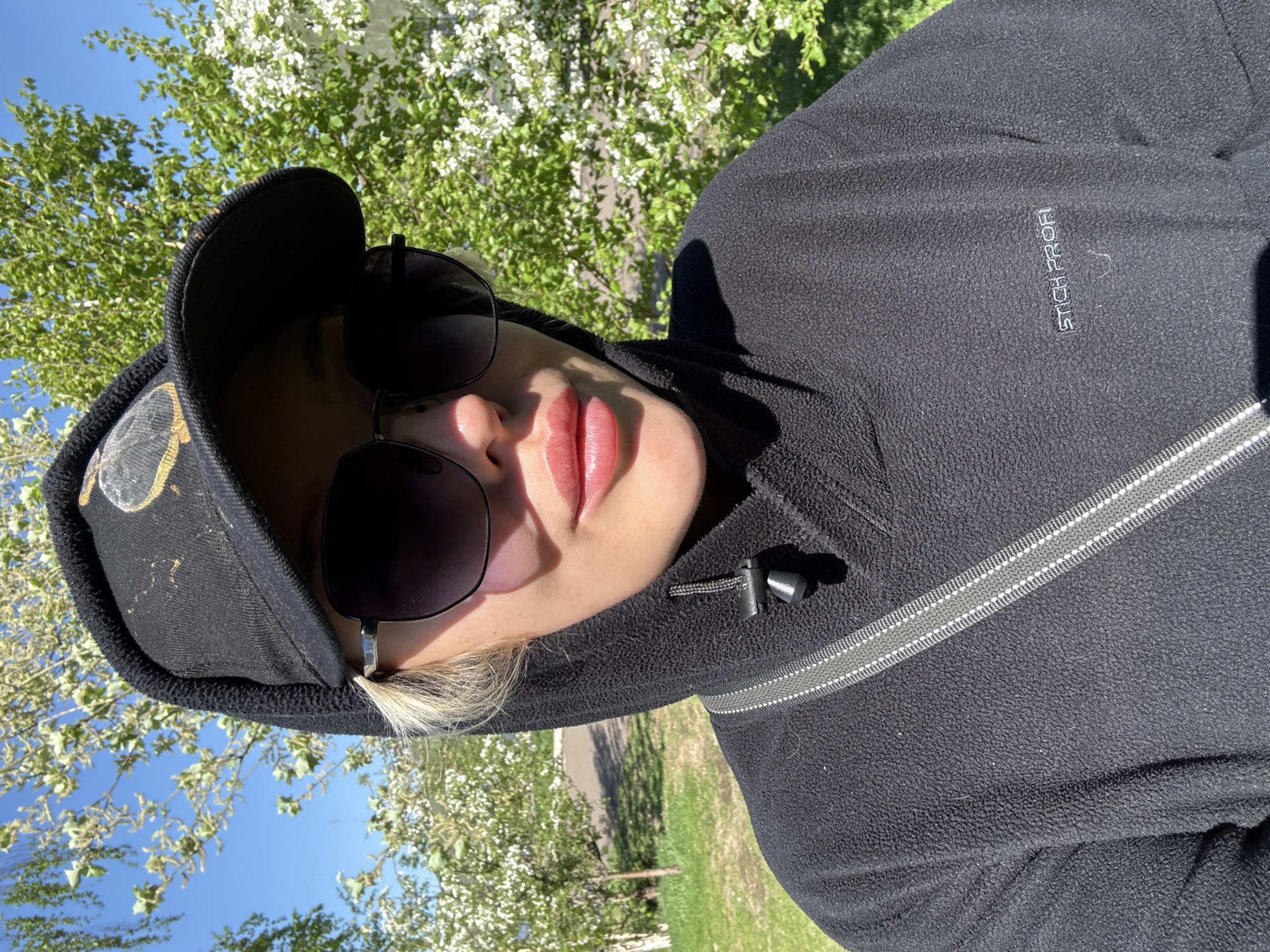 A woman wearing sunglasses and a black hooded fleece jacket is pictured from a close-up, low-angle perspective. She is also wearing a black cap with gold-colored designs on it. The jacket has a silver zipper and the words "STICH PROFI" embroidered on the right side. The background features green grass and trees with white blossoms against a blue sky. Without more information, the precise location within the city of Krasnoyarsk is not discernible.
