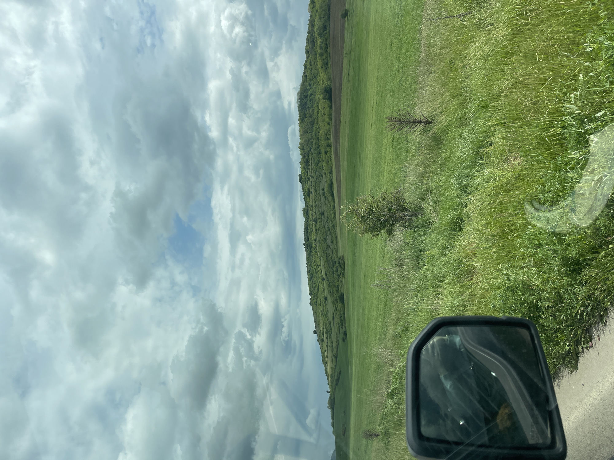The image depicts a landscape view from a vehicle, likely a car, moving along a road in what appears to be the rural or semi-rural outskirts of Batanovtsi, Bulgaria. The sky is filled with grey and white clouds with occasional patches of blue sky visible. A hill or slope is visible to the left of the frame, covered with green grass and some areas of forest. A field is located to the right, with tall green grass. The side mirror of the vehicle is visible in the lower right corner of the image, reflecting some objects that may be located within the vehicle. The road is visible to the right, made of asphalt or concrete. The overall environment suggests a countryside or suburban area in Batanovtsi, Bulgaria.