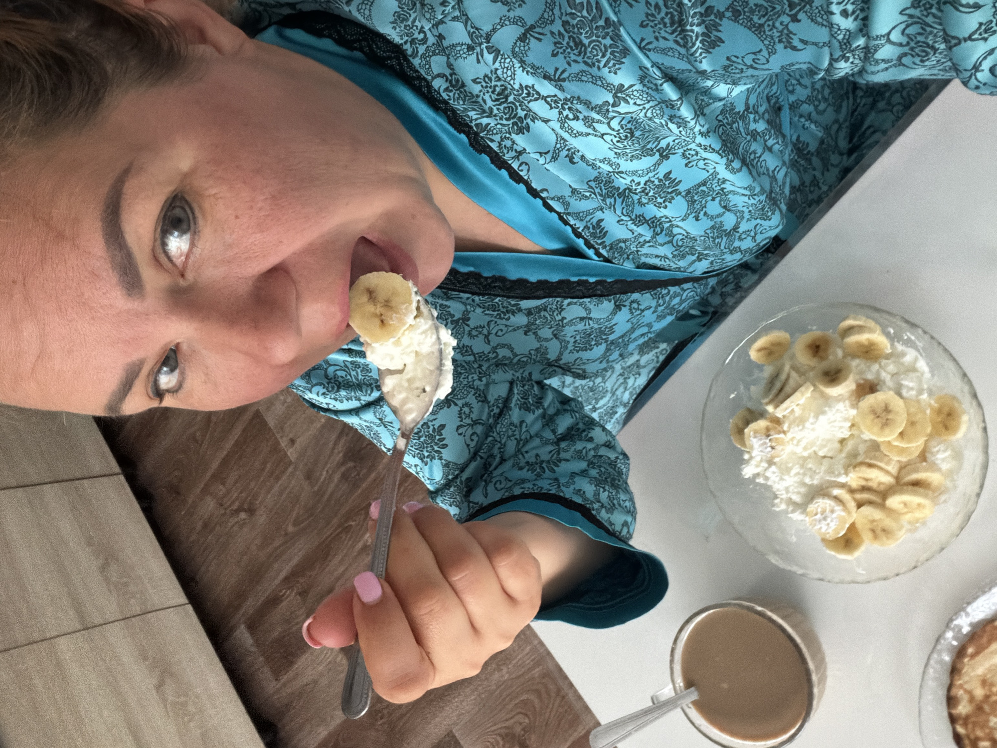 The image shows a woman eating a dessert of sliced bananas with cream from a bowl. She holds a spoon with a banana slice and cream on her tongue. She is wearing a blue robe with a floral pattern. A glass of coffee and pancakes are on the table next to the bowl. The scene appears to be in a private home. There is no evidence suggesting the location in the city of Leningradskaya, Russia.