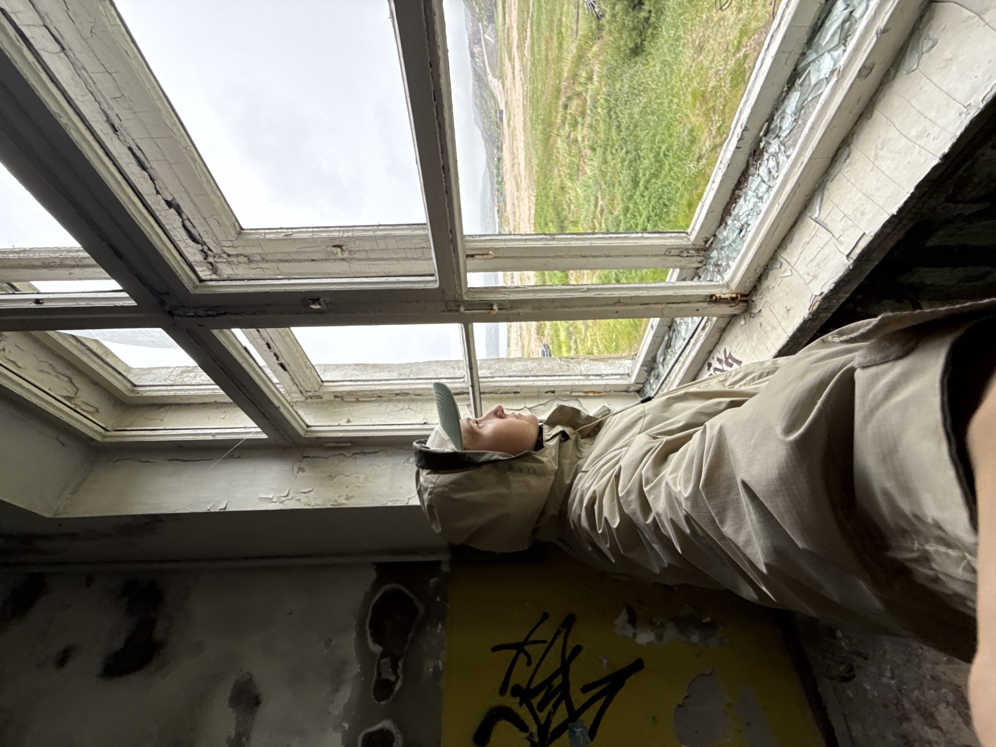 An individual wearing a tan hooded jacket and a light green baseball cap stands in a dilapidated room, looking out of a large, multi-paned window. The room's walls are distressed, with peeling paint and graffiti on the left side. The window frame is old and weathered, with cracks and chipped paint. The window itself is broken in places, with shards of glass scattered on the windowsill. Outside the window, a vast, green, and hilly landscape is visible under an overcast sky. The scene appears to be located in Teriberka, Russia, a remote coastal village known for its stark, abandoned industrial structures and dramatic natural scenery.