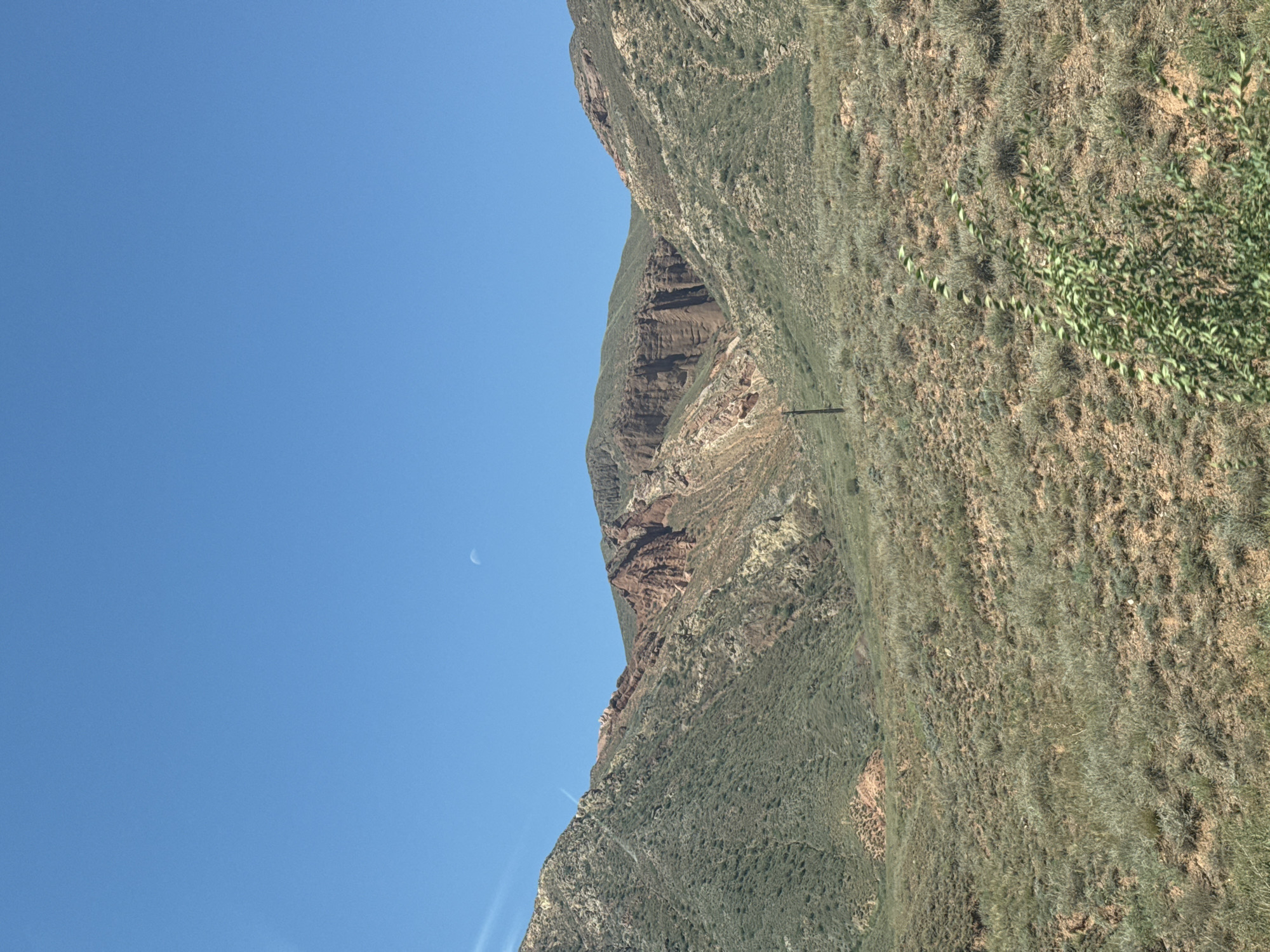 The image depicts a vast, arid mountain landscape under a clear, bright blue sky. The terrain is characterized by rolling hills covered in sparse, dry vegetation, predominantly consisting of grasses and low shrubs. The mountains themselves exhibit distinct geological formations, with exposed rock faces showing layers of red, brown, and yellow soil. In the distance, a sliver of the moon is visible against the sky. A single, dark utility pole stands in the mid-ground, a solitary man-made object in the natural environment. The overall impression is one of a remote, undeveloped, and rugged natural setting. There is no indication of human presence or activity beyond the utility pole.