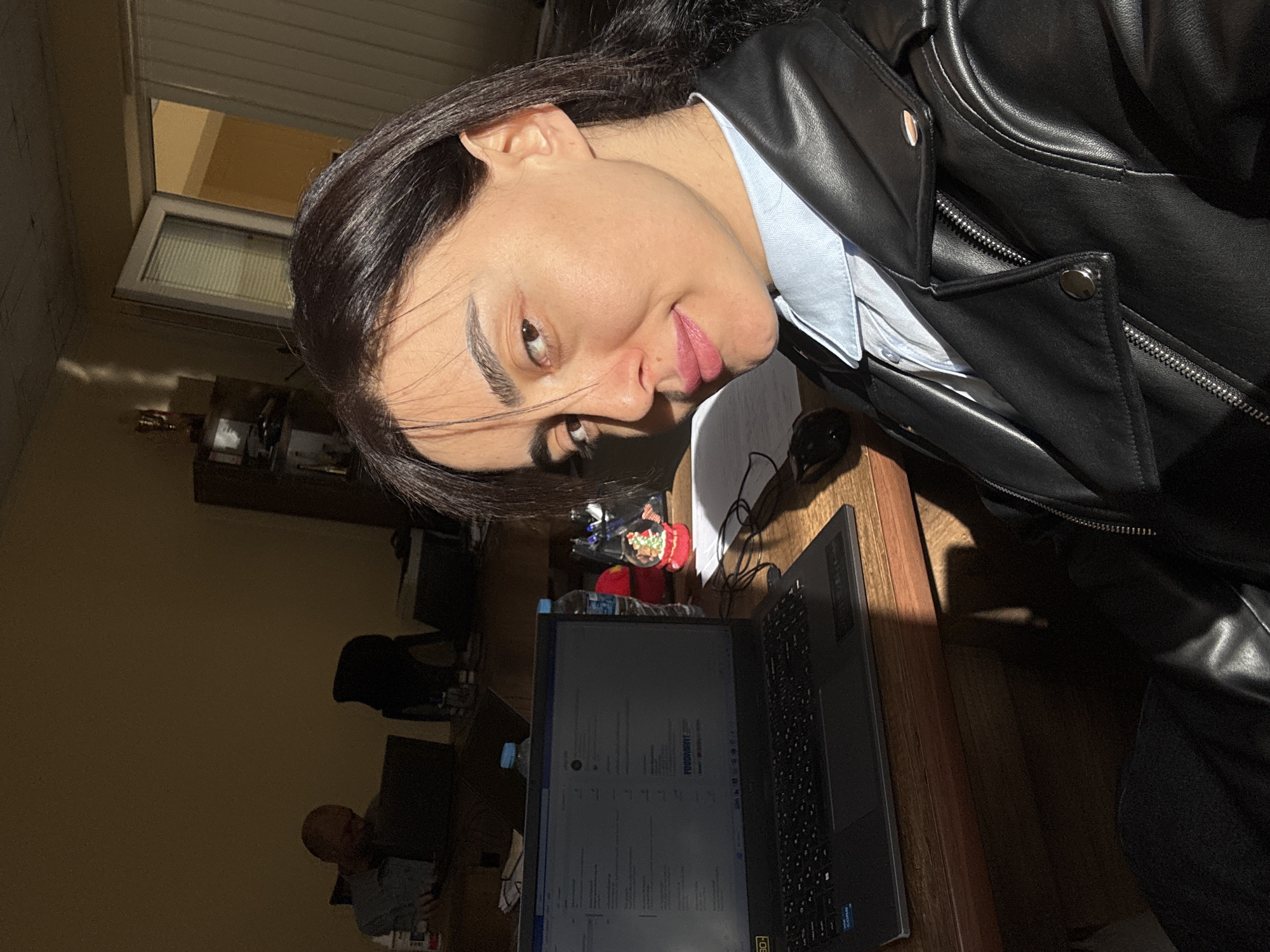 A woman with dark hair and brown eyes poses indoors, wearing a black leather jacket over a light blue collared shirt. She is positioned near a wooden desk with a black laptop displaying a webpage. The room is illuminated with natural light, possibly from a window visible in the background, which suggests an interior space. A male figure is visible in the background, seated at a desk. Additional objects include a water bottle, a Christmas ornament, and office supplies on the desk. The location is likely Tbilisi, Georgia.