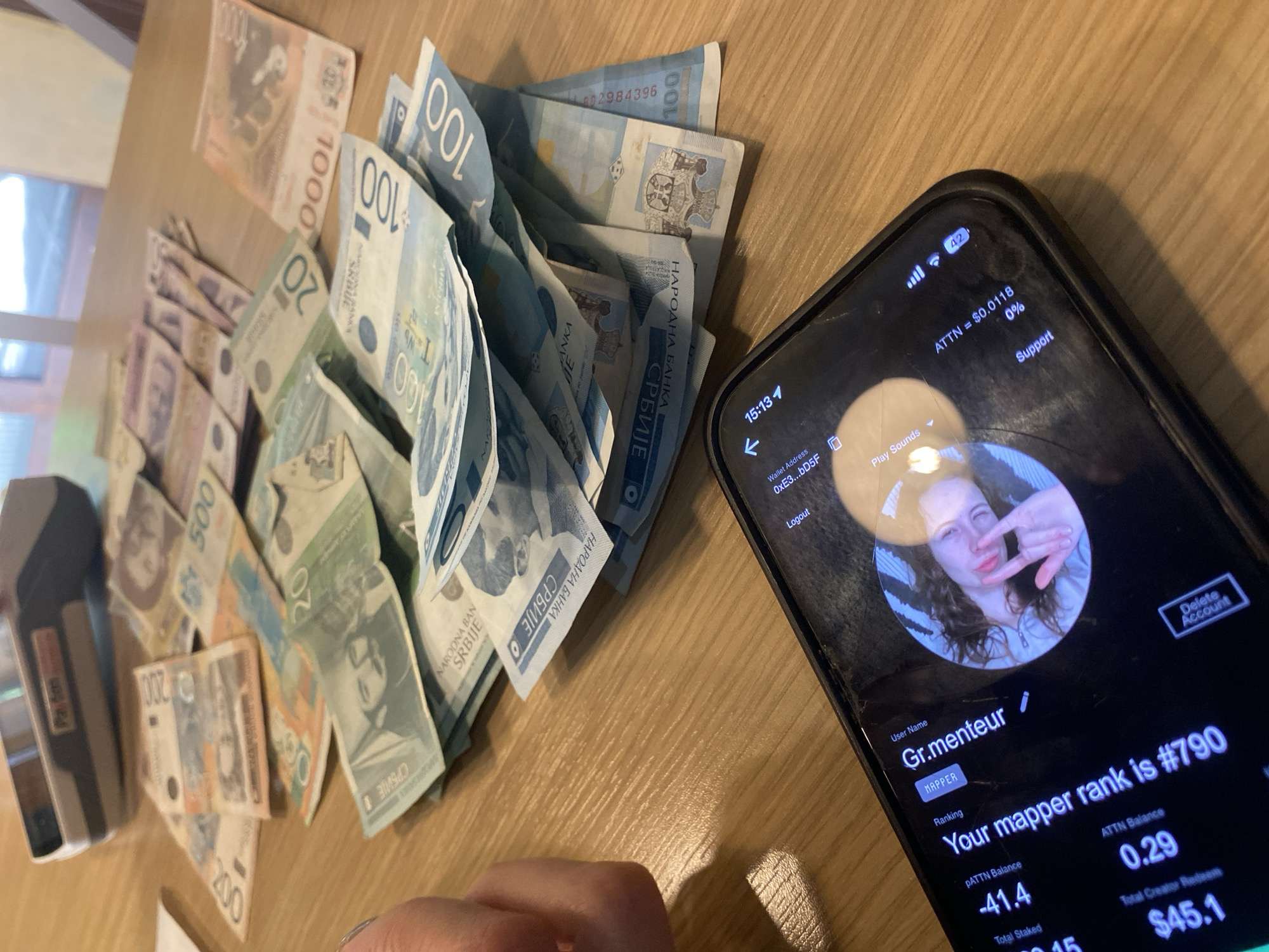 On a wooden table, there is a scattering of Serbian Dinar banknotes of various denominations (1000, 500, 200, 100, and 20). A black smartphone is placed next to the money, displaying the user profile "Gr.menteur" on an application. The application shows details like "Mapper rank is #790," "ATTN Balance 0.29," and "Total Creator Redeem $45.1." The phone screen also shows "ATTN = $0.0118" and indicates a "Support" option. A stapler is to the left of the money. A partially visible hand is present near the bottom of the image. The location, Novi Sad, Serbia, is inferred from the Serbian currency.