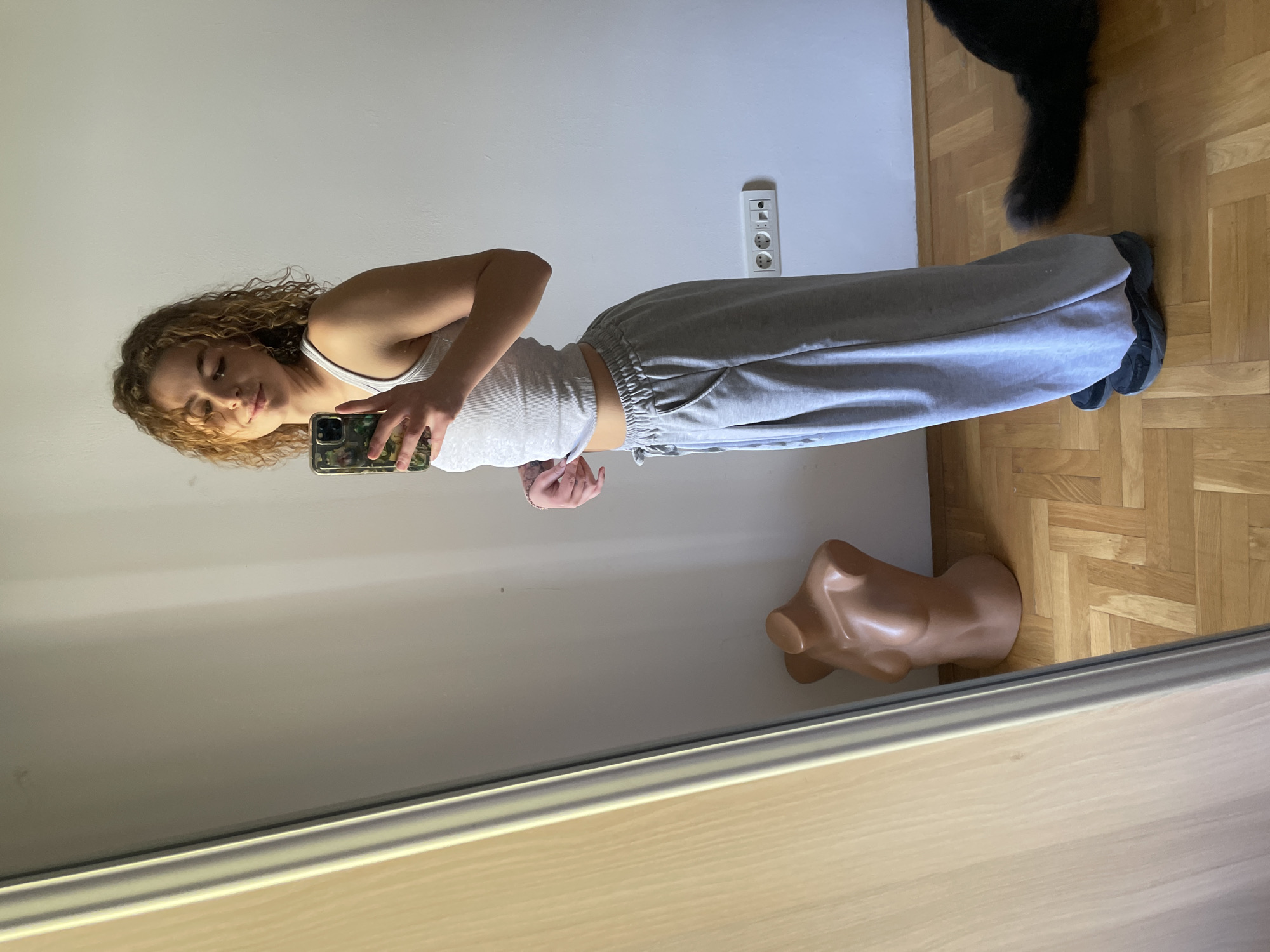 A woman with curly hair is taking a selfie in front of a mirror. She is wearing a white tank top and grey sweatpants. In the background, there is a wall with an electrical outlet, a wooden floor, a black object that appears to be a pet's tail, and a mannequin torso on the floor. The location in the city of Boreti, Montenegro, cannot be confirmed.