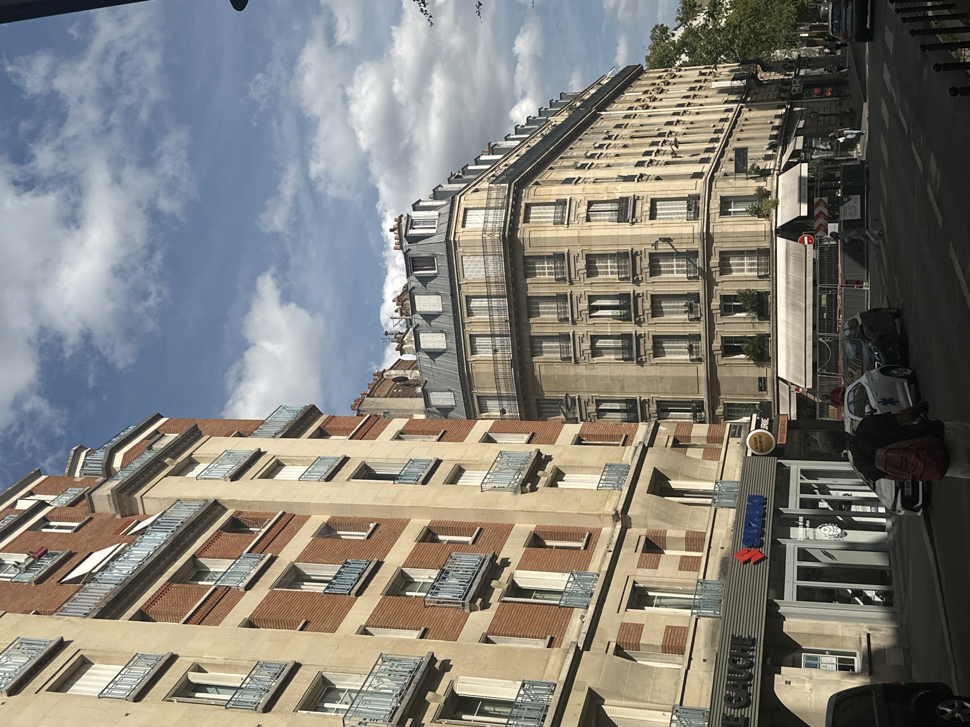 The image shows a street scene in Paris, France, with several buildings lining a road. The architecture features typical Parisian styles, with ornate balconies, mansard roofs, and stone facades. On the left, a red brick and cream-colored building with multiple stories and French balconies dominates the foreground. A Suzuki dealership is visible at the street level of this building, indicated by a prominent sign. Several cars are parked or driving on the street, and a few pedestrians are walking. The sky above is blue with scattered white clouds. To the right, a larger, lighter-colored building with many windows and balconies curves around a street corner. The overall atmosphere is that of a typical urban environment in Paris on a clear day.