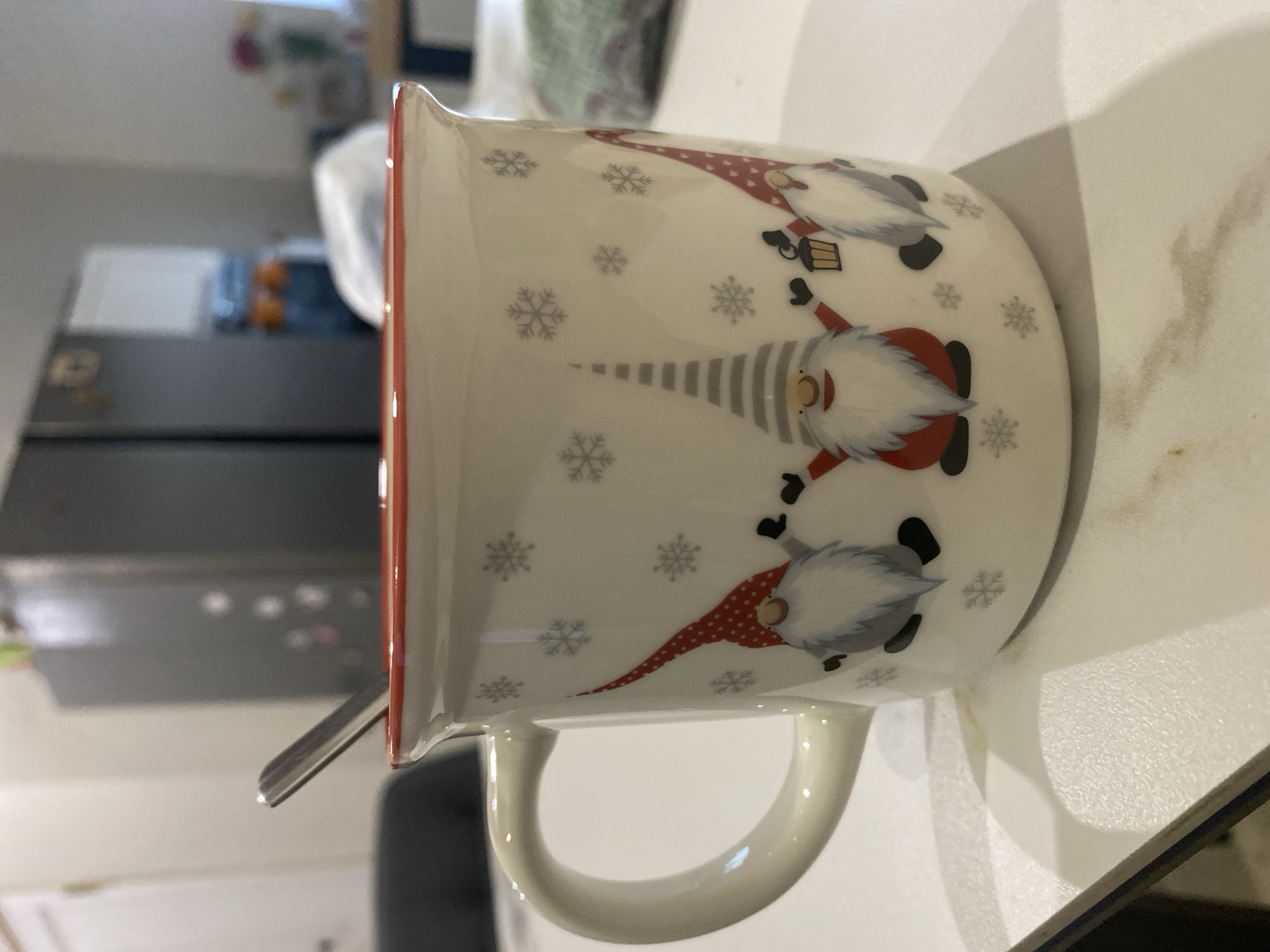 A close-up of a white ceramic mug with a red trim and a winter gnome motif, placed on a white countertop. The mug features three gnomes in different hats and poses, interspersed with snowflake designs. A silver spoon is inserted into the mug. The background shows a partial view of a kitchen with a stainless steel refrigerator. There is no explicit information in the image to confirm the location as the city of Chaikovsky, Russia.