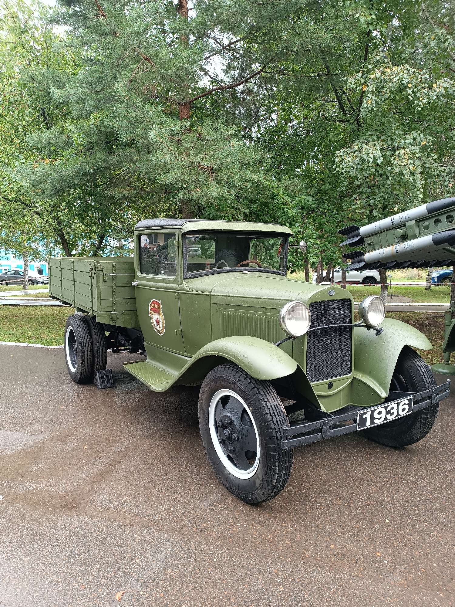 A vintage, olive-drab truck, manufactured in 1936, is parked outdoors on a wet asphalt surface. The truck has a wooden cargo bed and a distinctive red and gold emblem on its side. Behind the truck, a multiple rocket launcher system is visible, suggesting the location may be a memorial or exhibition related to military history. The background features lush green trees and a cloudy sky. The truck is the primary subject, with its historical significance and the contrast between the civilian vehicle and military equipment being notable elements. The scene appears to be in a public space in the city of Chaikovsky, Russia, possibly a park or a museum grounds.