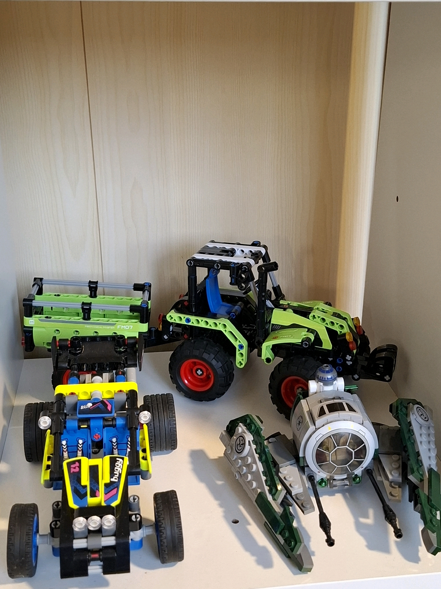 The image shows a close-up, eye-level, interior shot of a white shelf with a light wood-grain textured background. The shelf is holding several Lego construction toys. In the foreground, on the left, is a blue and yellow Lego buggy with black wheels and detailing. To its right and slightly behind it is a green and black Lego tractor with large red and black wheels. In the background, on the left, is a green Lego trailer with grey bars and black connectors, attached to a black Lego frame. To the right of the tractor and in the foreground is a grey and green Lego spaceship with a round cockpit and two protruding arms with green accents. The lighting appears to be natural, coming from the right, casting shadows to the left of the objects. There are no people or animals present. The scene is static and does not depict any actions or interactions. The location is not specified, and there is no indication it is in the city of Tyumen, Russia.