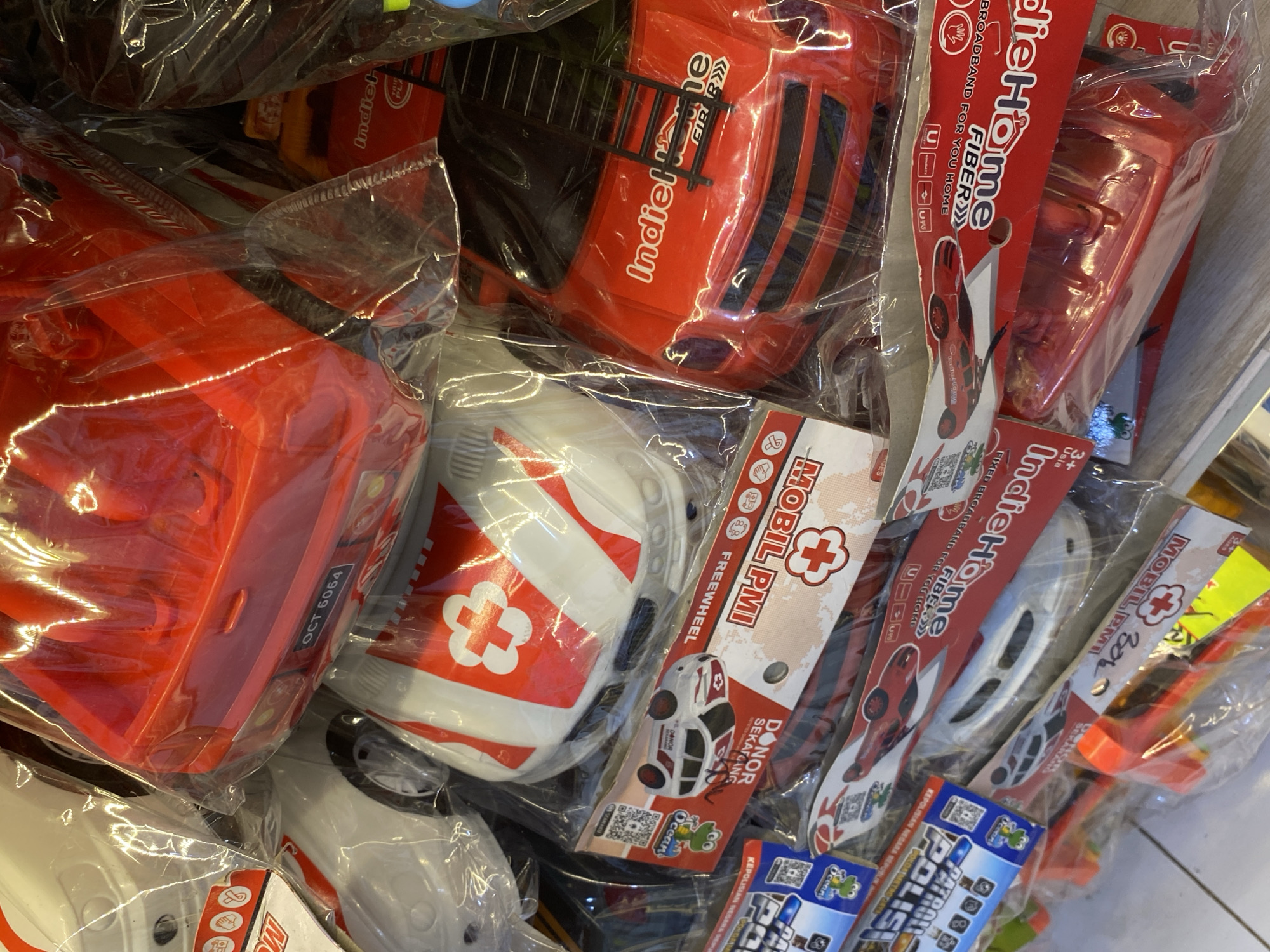 The image shows a close-up, eye-level view of a stack of toy vehicles, likely fire trucks and ambulances, packaged in plastic and cardboard. The toys are predominantly red and white, with some black accents. Several labels are visible on the packaging, some featuring red crosses and text in what appears to be Indonesian, such as "MOBILE PMI" and "DONOR SEKARANG". One label shows a license plate with "OCT 6064". The toys are arranged in a retail setting, possibly a shelf or display in a store. The floor tiles are visible in the lower portion of the image. The environment appears to be indoors, with artificial lighting. No people are visible in the image.