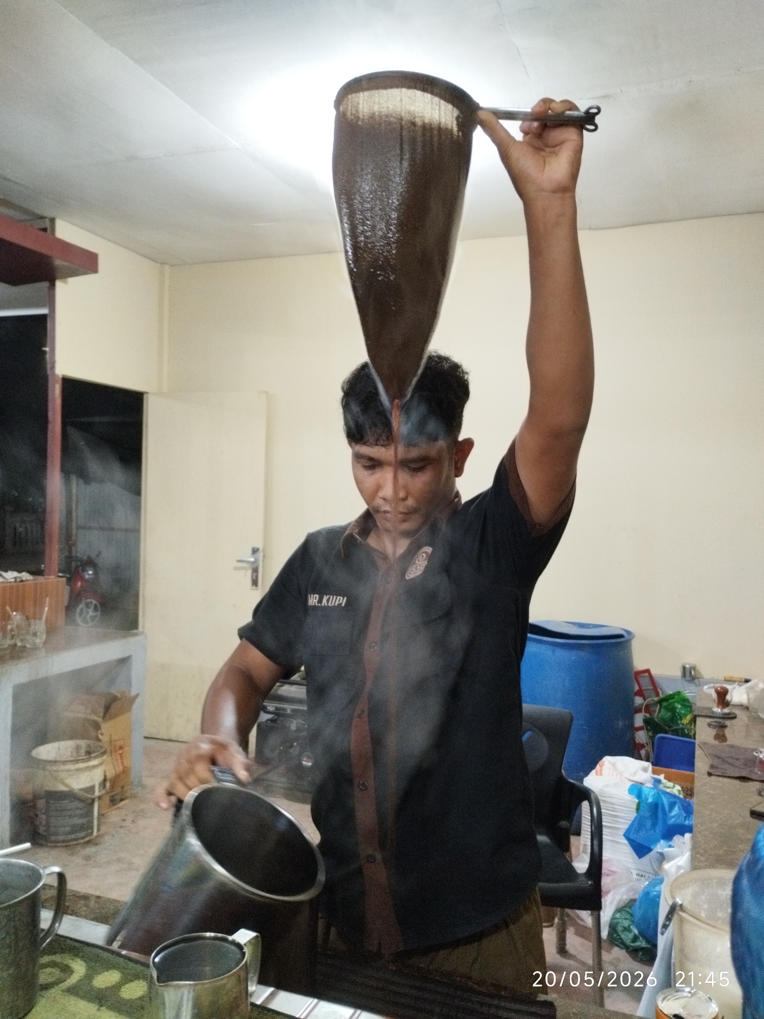 A man stands indoors, likely in a food service establishment, performing a coffee-making process. He is holding a sock-like strainer filled with coffee grounds high in his right hand, above his head, as liquid coffee filters down. In his left hand, he holds a metal pitcher to catch the coffee. Steam rises from the coffee. The man wears a dark-colored shirt with the name "MR. KUPI" embroidered on the chest. The background includes a counter with metal mugs, a blue plastic barrel, and various containers. A door and an open space leading to a dimly lit area are visible. The date "20/05/2026 21:45" is stamped on the lower right of the image. The location is Lhokseumawe, Indonesia, based on the local coffee-making methods depicted.