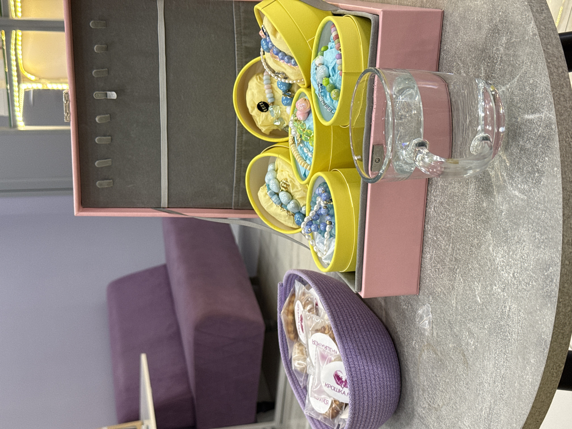 The image depicts an indoor scene, likely a retail or personal storage space. In the foreground, a grey, textured round table holds a clear glass mug filled with water and a crumpled piece of clear plastic. Behind the table, a pink jewelry box is open, revealing several yellow cylindrical containers filled with colorful beaded bracelets. To the left of the box, a lavender braided basket contains individually wrapped food items. In the background, a purple upholstered sofa is visible against a plain, light purple wall. The overall impression is one of organized storage and display. The image does not contain any identifiable landmarks or contextual clues that would place it specifically in Lyubertsy, Russia.