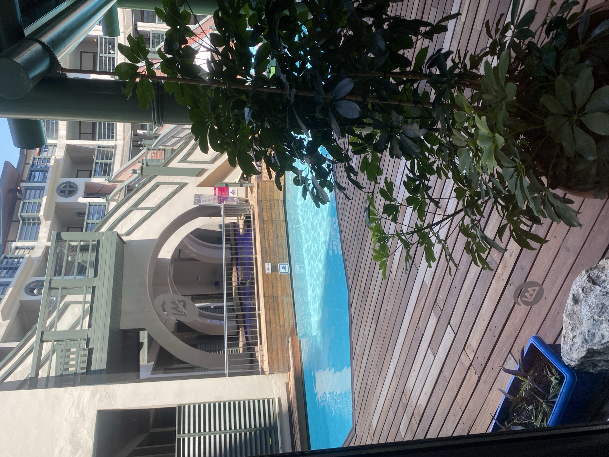 The image shows a swimming pool area, likely at a resort or hotel, with a wooden deck in the foreground. The pool is bright blue and reflects the sunlight. A large potted plant with dark green leaves is in the lower right corner of the frame, partially obscuring the view. In the background, a multi-story building with balconies and staircases is visible. The architecture suggests a modern design with touches of Mediterranean or tropical influences. Signs indicating the depth of the pool (1.40 m) and a diving symbol are present near the pool's edge. There is no indication of the city of Aksu, Turkey in the image itself.