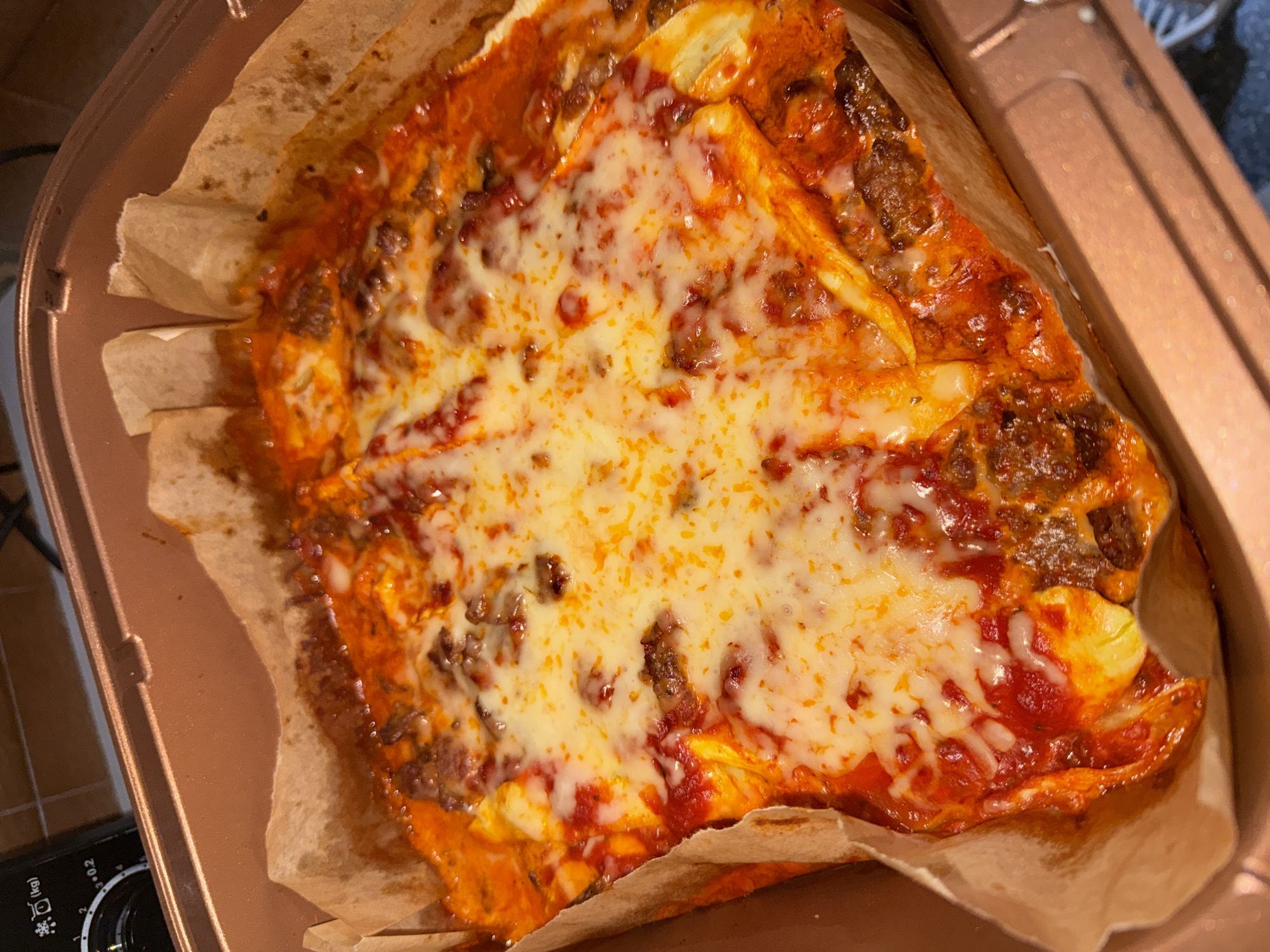 A close-up shot depicts a portion of lasagna in a metallic, square baking pan lined with parchment paper. The lasagna is covered in melted cheese, with visible layers of pasta, tomato sauce, and ground meat. The pan rests on a stovetop, with a knob and settings visible at the lower left corner. The scene appears to be indoors, potentially in a home kitchen setting. There is no indication of the city of Burgas, Bulgaria or any people.