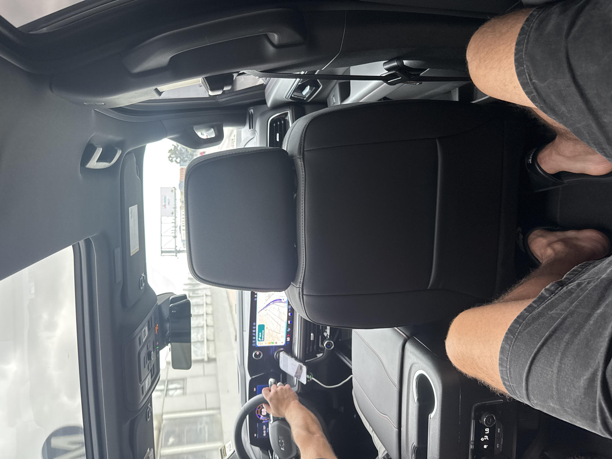 An interior view from the backseat of a moving vehicle on a highway in Los Angeles, California, shows the driver's hands on the steering wheel and a smartphone mounted on the dashboard. The driver is wearing a dark shirt. The front passenger seat is empty. In the foreground, a person's legs are visible, wearing dark denim shorts and black flip-flops. The vehicle's interior includes a sunroof, a rear-view mirror, and climate control buttons. Outside the car, a highway overpass and distant buildings are visible.