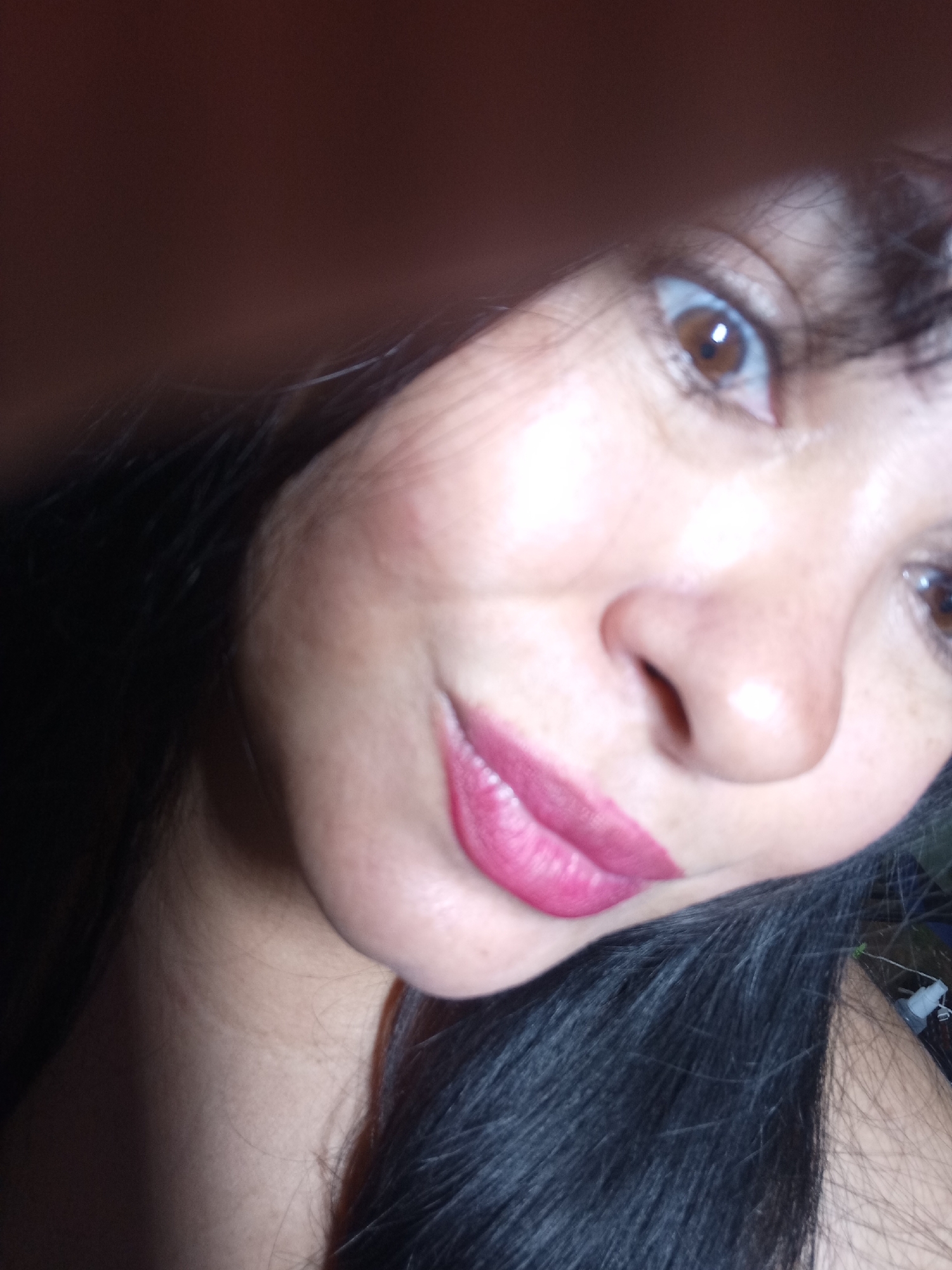 A close-up selfie captures a person's face from a low, tilted angle. The subject, likely a woman, has dark hair that frames her face and extends into the bottom right corner of the frame. Her skin tone appears warm. She has wide, brown eyes, with the right eye looking directly at the camera, while the left eye is partially obscured by something dark in the upper left foreground, possibly a hand or hair. Her lips are painted with a vibrant fuchsia or magenta lipstick, and she has a subtle smirk or soft smile. Her nose is visible from a slight angle, and there is a natural sheen on her skin, particularly on her nose and cheek. The lighting is bright on her face, suggesting a flash or direct light source, while the background is mostly dark and out of focus. In the very bottom right corner, a faint, blurred white object, possibly a small bottle or dispenser, can be barely seen against a dark background. No visible text is present.