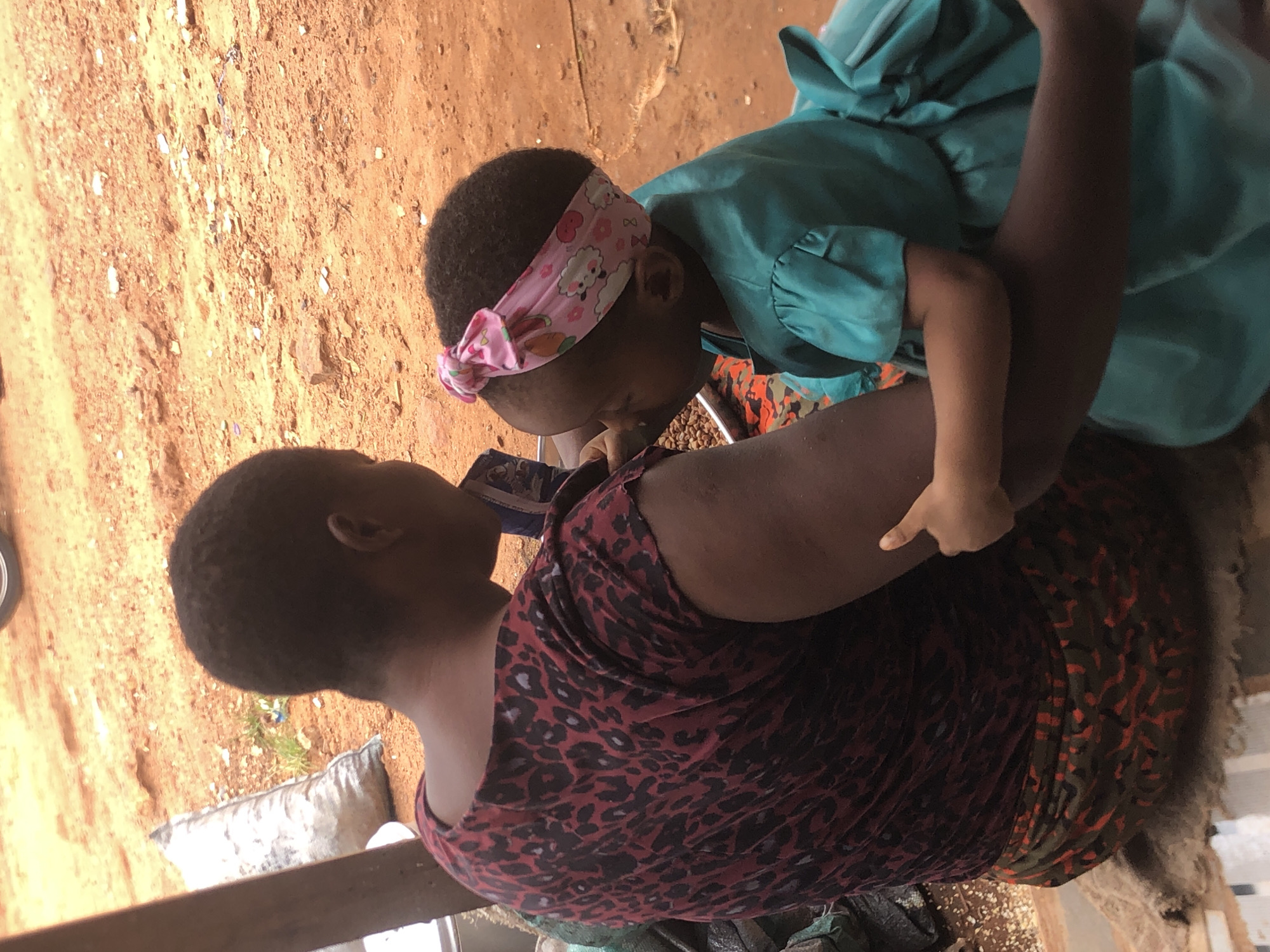 The image captures an intimate moment between an adult and a young child in an outdoor or open-air setting in Ukwu-Abwa, Nigeria. The ground is a reddish-brown dirt or sand, scattered with small white and brown debris.
The subjects are an adult, likely a woman, and a young child. The adult, seen from the back and side, has dark skin and short, dark hair, and wears a red and black leopard print sleeveless top with a multi-patterned skirt featuring red and green hues. The child, also with dark skin and short, dark curly hair, is wearing a light blue or teal dress and a pink headband adorned with cartoon sheep and hearts.
The child is leaning into the adult, with their head bent down. A metal bowl containing small, dark, granular items, possibly food like beans or coffee beans, is visible between them. This suggests the adult is feeding the child or they are sharing a meal. The child's left arm is bent, with a hand visible, appearing to rest on the adult's side or arm. The adult is looking downwards towards the child or the activity.
The bright, direct lighting indicates it is daytime. The overall scene conveys a tender moment of care or bonding. No visible text is present in the image. The rustic setting with the dirt ground provides a contextual clue to the location.