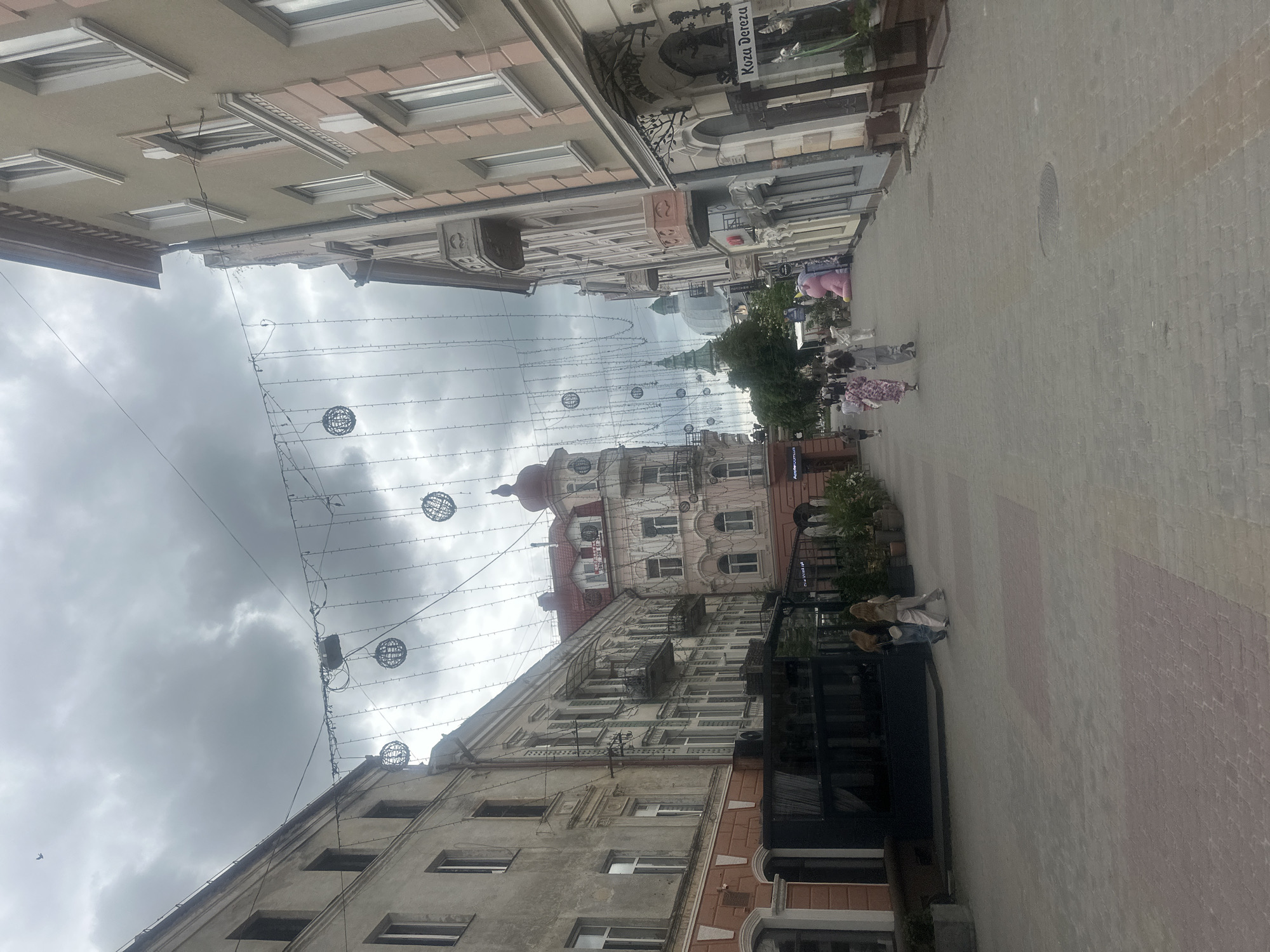 The image depicts a street scene in Ternopil, Ukraine, likely a pedestrian area lined with historic buildings and decorated with string lights and spherical ornaments. In the foreground, a street paved with interlocking bricks leads into the distance. Two women are walking away from the camera on the left side of the street. Further down the street, other pedestrians are visible, as well as shopfronts and outdoor seating areas. A large pink inflatable object, resembling a pig, is situated on the right side of the street. The architecture of the buildings suggests an older European city, with ornate facades and balconies. The sky is overcast, with dark, heavy clouds. In the distance, a green dome topped with a spire is visible, likely part of a church or administrative building. A sign on the right side of the street reads "Koza Dereza."