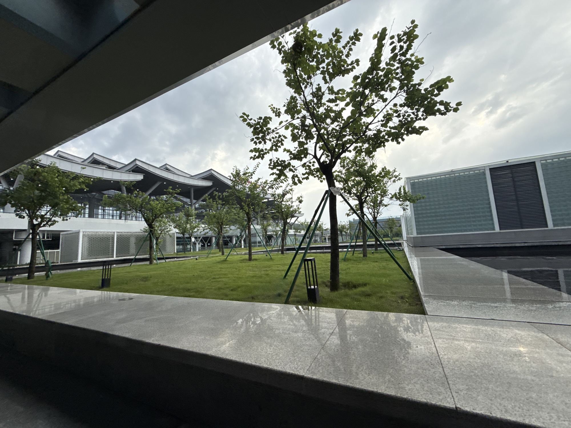The image shows a modern architectural complex with a green lawn and trees. The building features a distinctive, undulating roofline, with multiple sections that appear to be layered and curved. The facade is characterized by large windows and horizontal louvers. The grounds are landscaped with a well-maintained lawn, interspersed with young trees that are supported by green stakes. Several black, rectangular light fixtures are visible on the lawn. In the foreground, a grey, granite-like surface is visible, reflecting the sky and surrounding structures. The sky is overcast with a mix of grey clouds. This scene is located at the Huizhou High-Speed Railway Station in Huizhou, China.