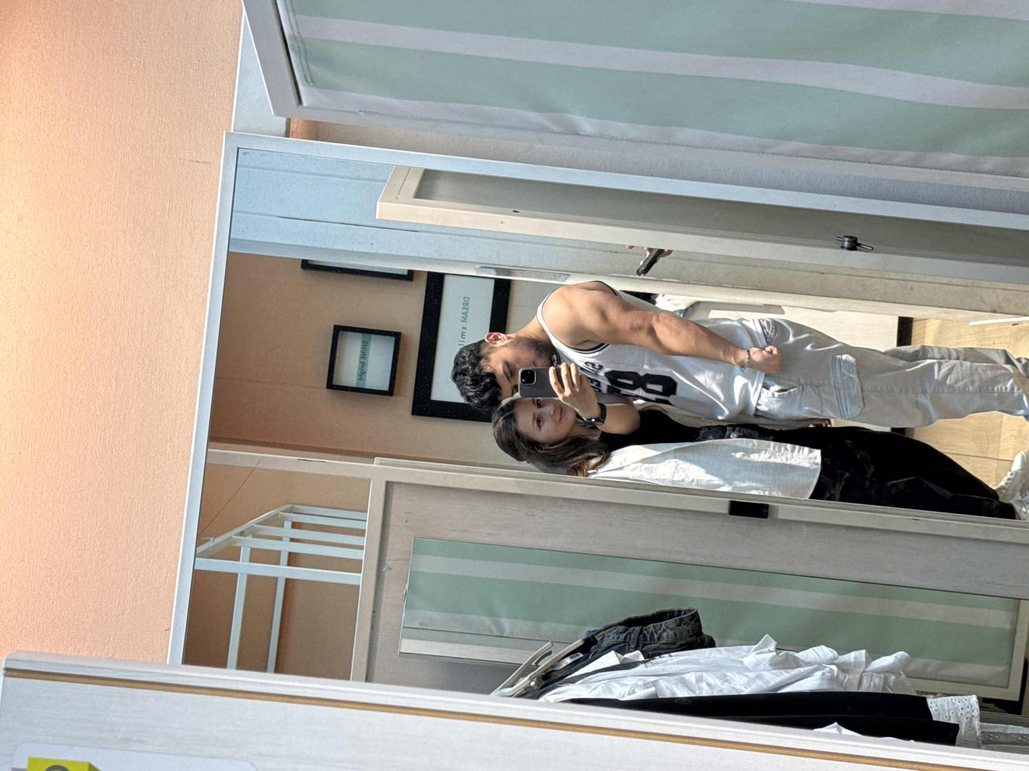 The primary image is a mirror selfie featuring a couple, presented rotated 90 degrees counter-clockwise. In its intended upright orientation, the photograph captures a man and a woman reflected in a large, vertical mirror within an indoor setting, likely a dressing room or a room with a prominent mirror.
The man, standing behind the woman, has dark hair and a beard, and wears a white sleeveless top with the number '88' or similar prominently displayed, paired with light-colored trousers. He appears muscular. The woman, with long brown hair, is holding a dark-colored smartphone to capture the mirror image and is wearing a black top. She is looking towards the phone with a slight smile, conveying a relaxed and happy demeanor.
The background reflected in the mirror reveals a light peach-colored textured wall. On this wall, two framed pictures are visible. One black frame contains mirrored text that appears to be "DREAM IS A MAJOR" (likely part of an original phrase like "DREAM IS A MAJOR KEY"). Below it, another black frame displays mirrored Cyrillic script. The presence of Cyrillic text is a significant location cue, consistent with the specified context of Varna, Bulgaria.
Above the couple's reflection, a light mint-green and white striped awning or blind is visible. In the foreground reflection, below the couple, various clothing items are casually placed, including a dark denim-like garment and white shirts. The scene is brightly and evenly lit, suggesting it is daytime.