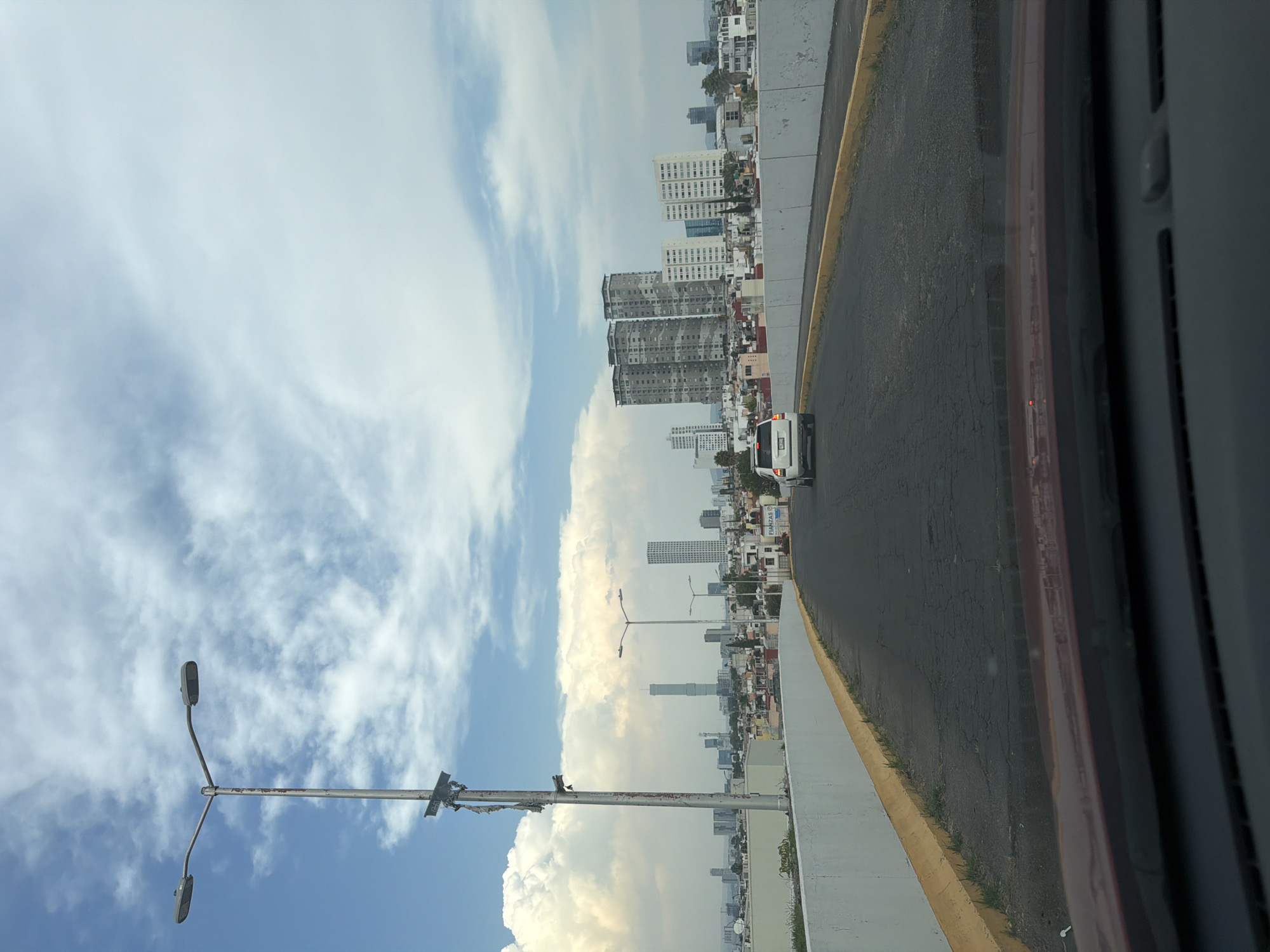 The image shows an urban street scene in San Bernardino Tlaxcalancingo, Mexico, as viewed from a moving vehicle. The street is asphalt with a concrete shoulder. In the background are multiple buildings with light colored exteriors. A white vehicle is visible on the road ahead. Streetlights are along the roadside. The sky is blue with clouds. The interior of the vehicle is visible in the foreground.