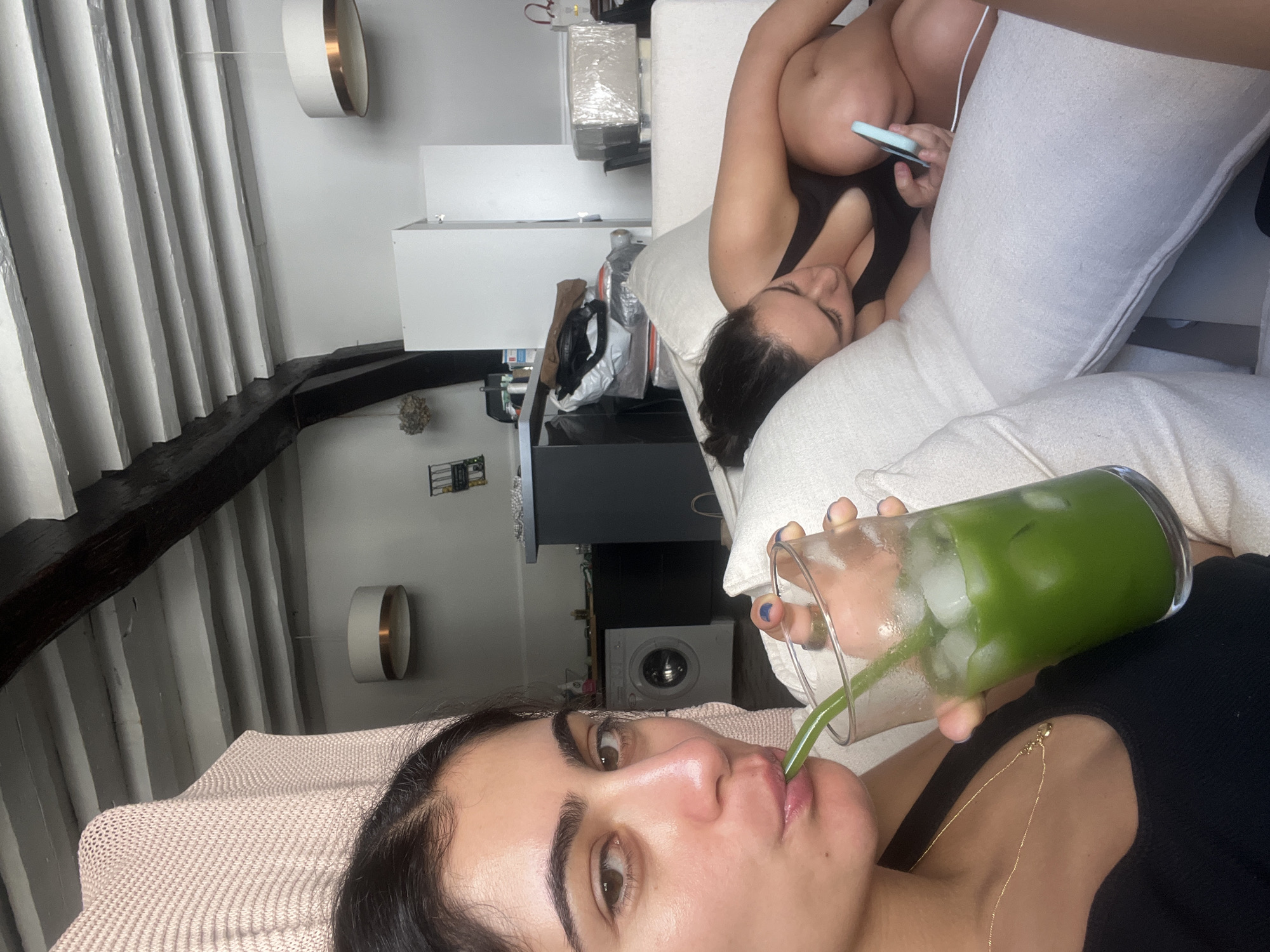 The media file shows two people indoors, likely in a living room. The foreground features a person drinking a green beverage with ice from a clear glass using a green straw. Their face is partially visible, with dark eyebrows, brown eyes, and pink lips. They are wearing a black top and a necklace.
In the background, another person is lying on a sofa, holding a light blue smartphone. They are wearing a black top and their body is partially visible. The sofa is light-colored, with several large cushions.
The room appears to have slanted ceilings with exposed wooden beams, suggesting an older building, possibly an attic space. There are white walls and some decorative items visible, including a cylindrical light fixture with a copper band on the wall. Furniture includes a black cabinet with shelves and a washing machine.
The lighting suggests it could be daytime, as there is ambient light coming from the windows, although the exact time of day is not discernible. The scene conveys a relaxed, casual atmosphere. There is no visible text in the media file. The location is specified as Paris, France.